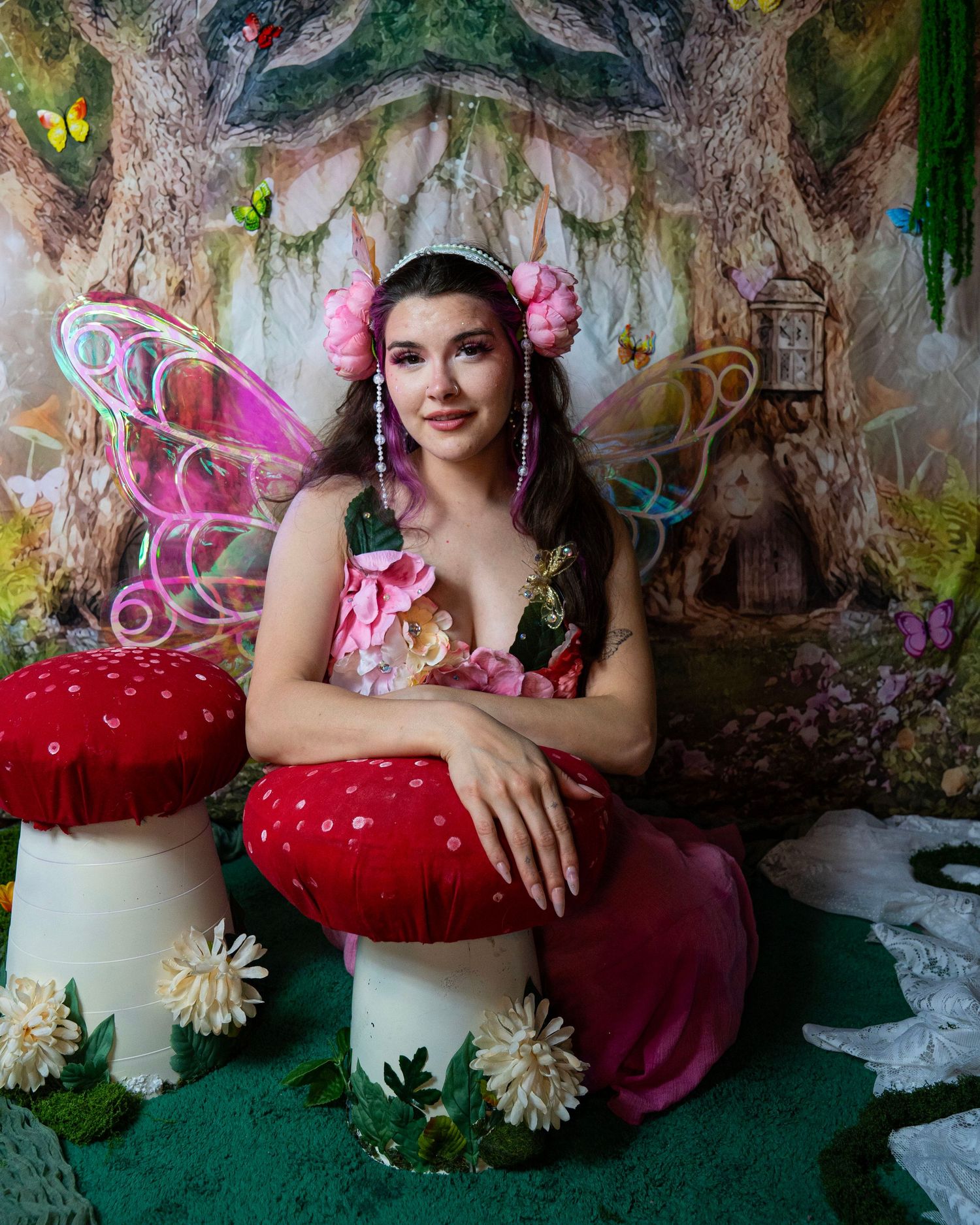 Brunette woman in fairy outfit with rainbow wings and pink flower crown sits beside red mushrooms in forest. Fantasy Photoshoot