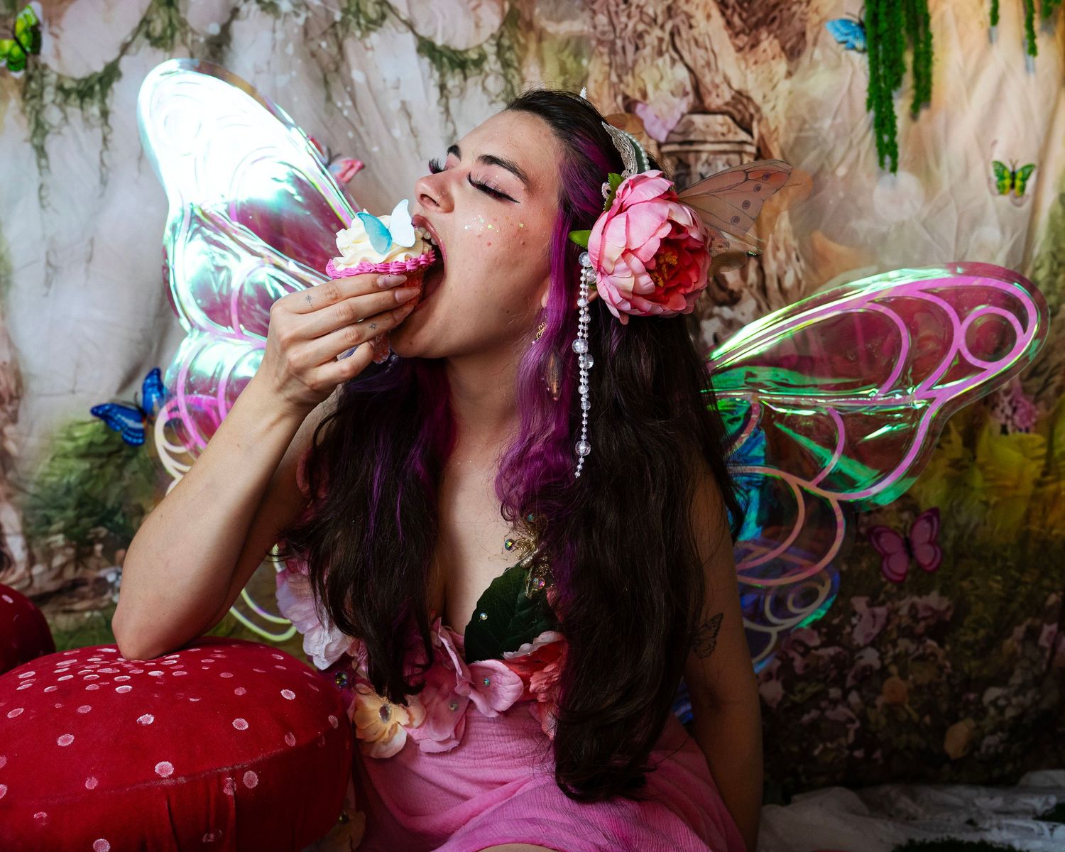 Woman in pink fairy costume with colorful wings eats cupcake in enchanted forest photography studio setting. Fantasy Photoshoot