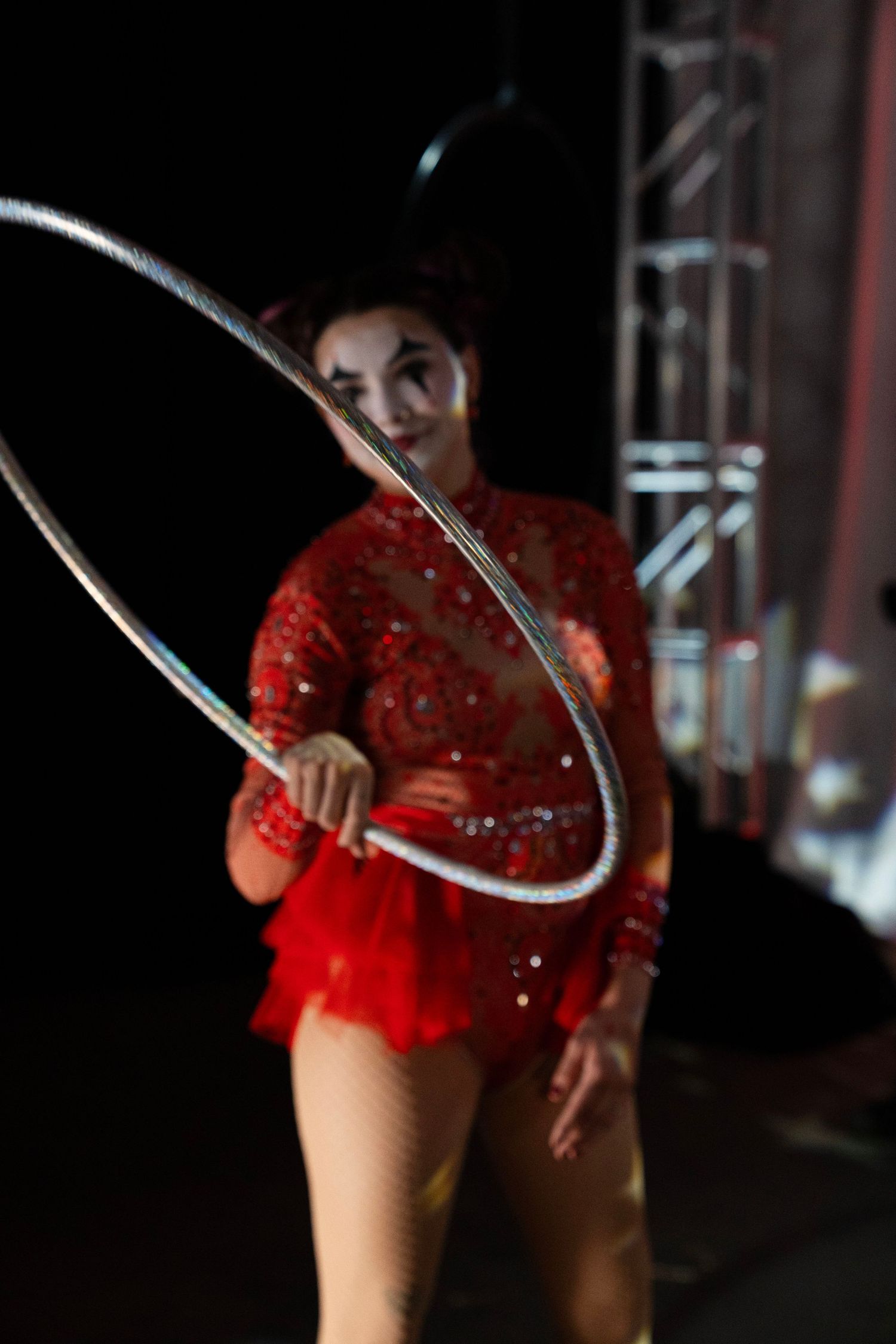 Circus performer in red sequin leotard spins a silver hula hoop on a dark stage.