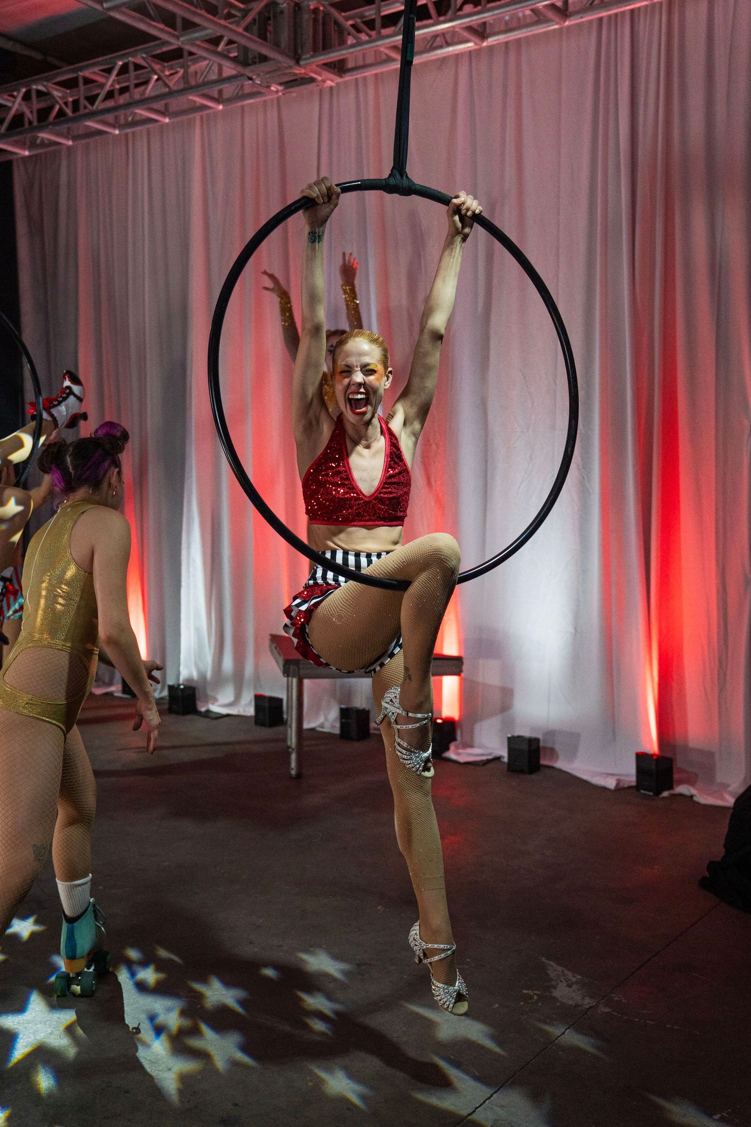 Smiling aerialist in red sequin costume performs on aerial hoop at a circus-themed stage performance. Circus Photoshoot