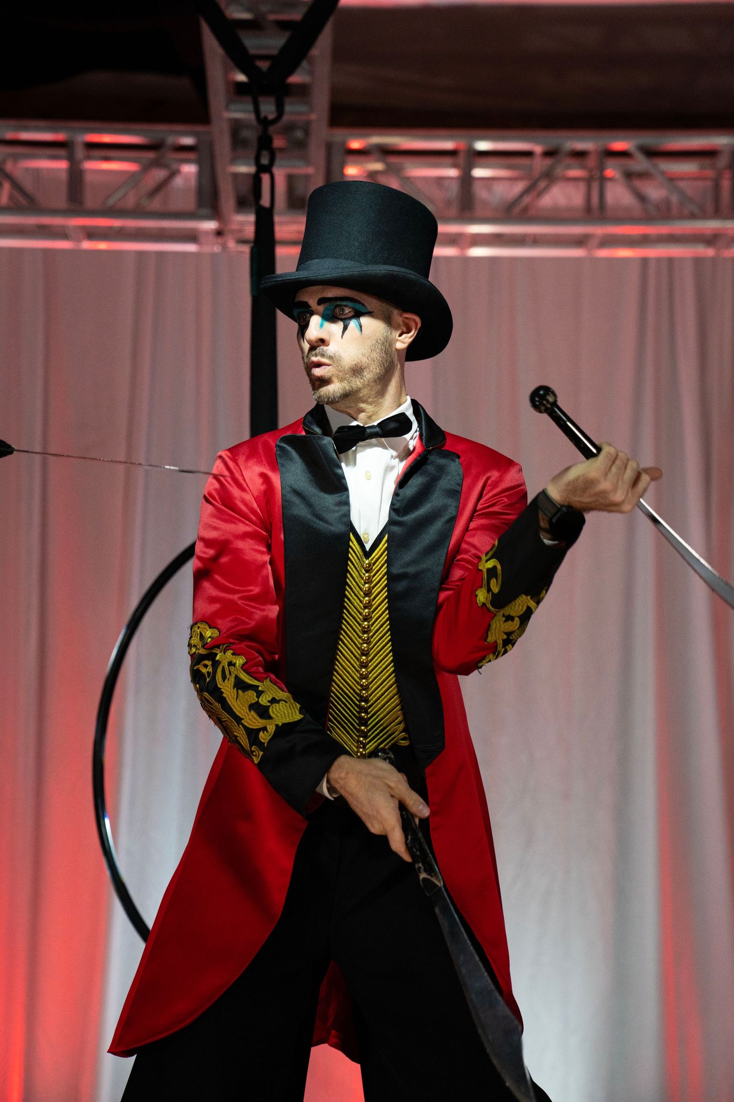 Circus ringmaster in red tailcoat and top hat poses with a sword and whip on a performance stage.