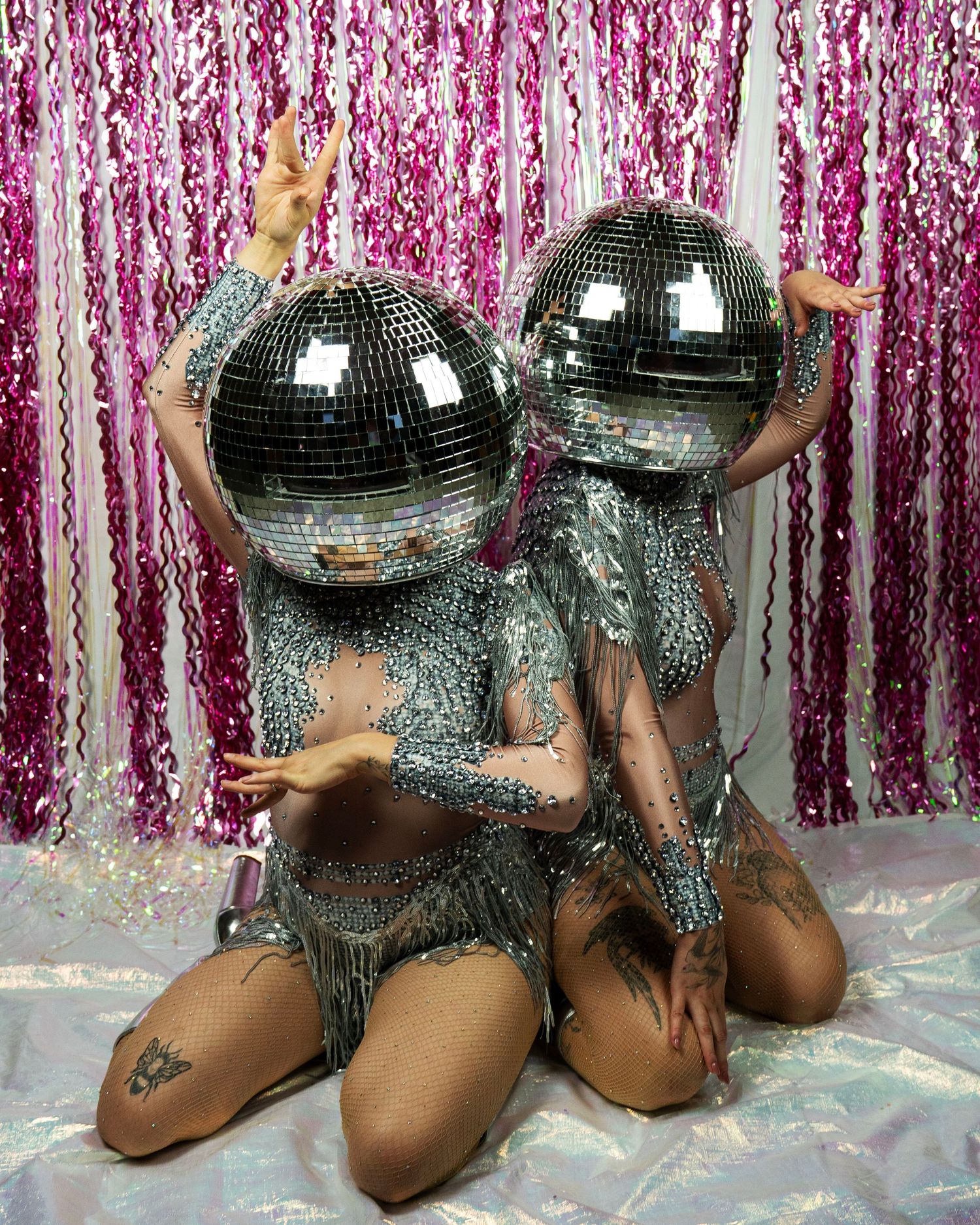 Two disco ball-headed performers in silver rhinestone fringe costumes pose kneeling on iridescent floor.