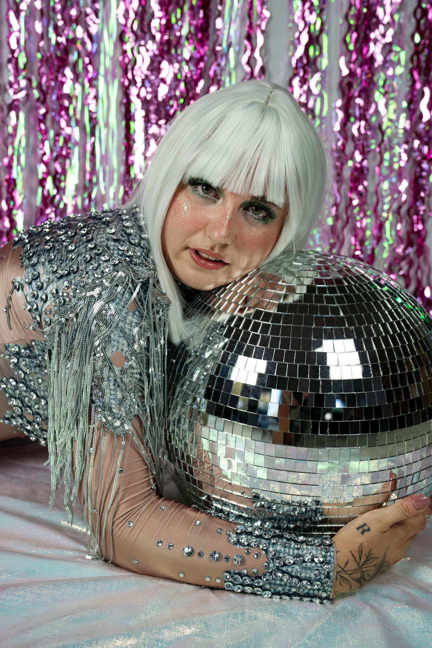 Performer in silver fringe sequin top with white wig reclines against disco mirror ball on white reflective surface.