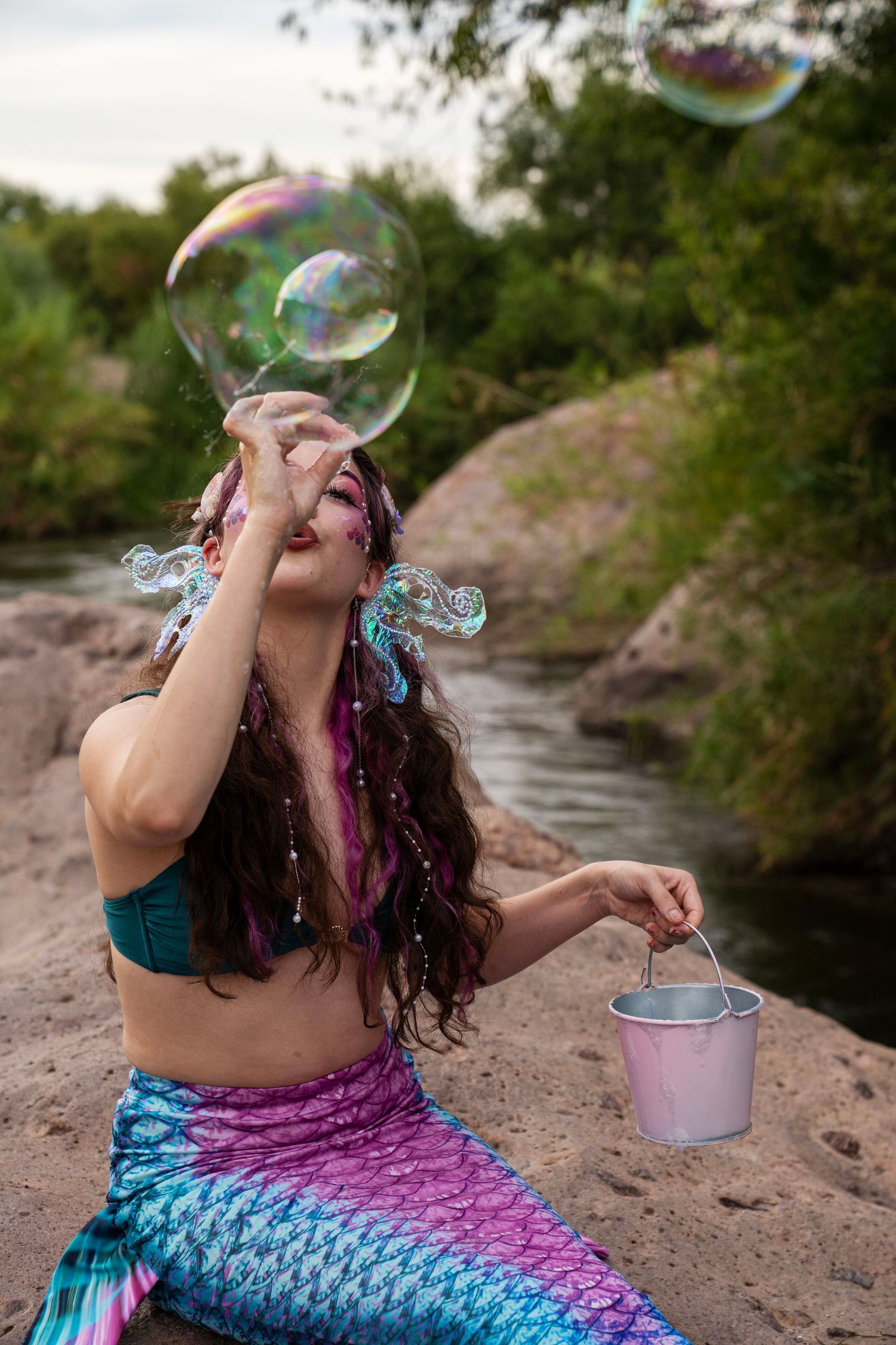 Woman dressed as a mermaid laughing while touching a large floating soap bubble near a river. Fantasy Photoshoot