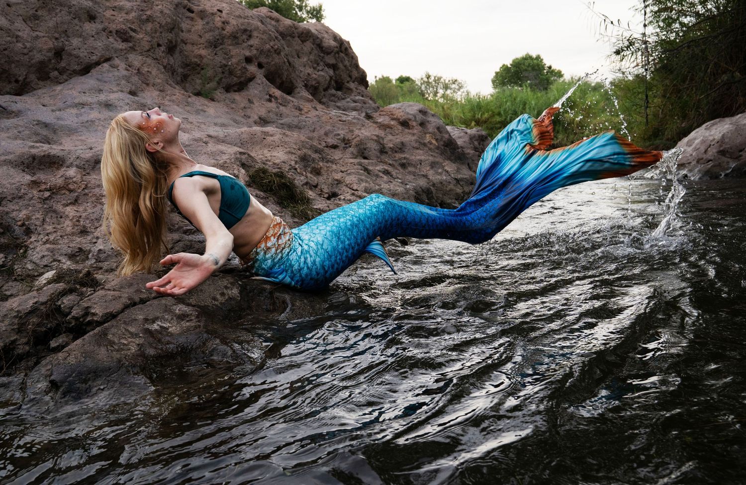 A woman in a shimmering blue mermaid tail reclines gracefully on wet rocks beside a river. Fantasy Photoshoot