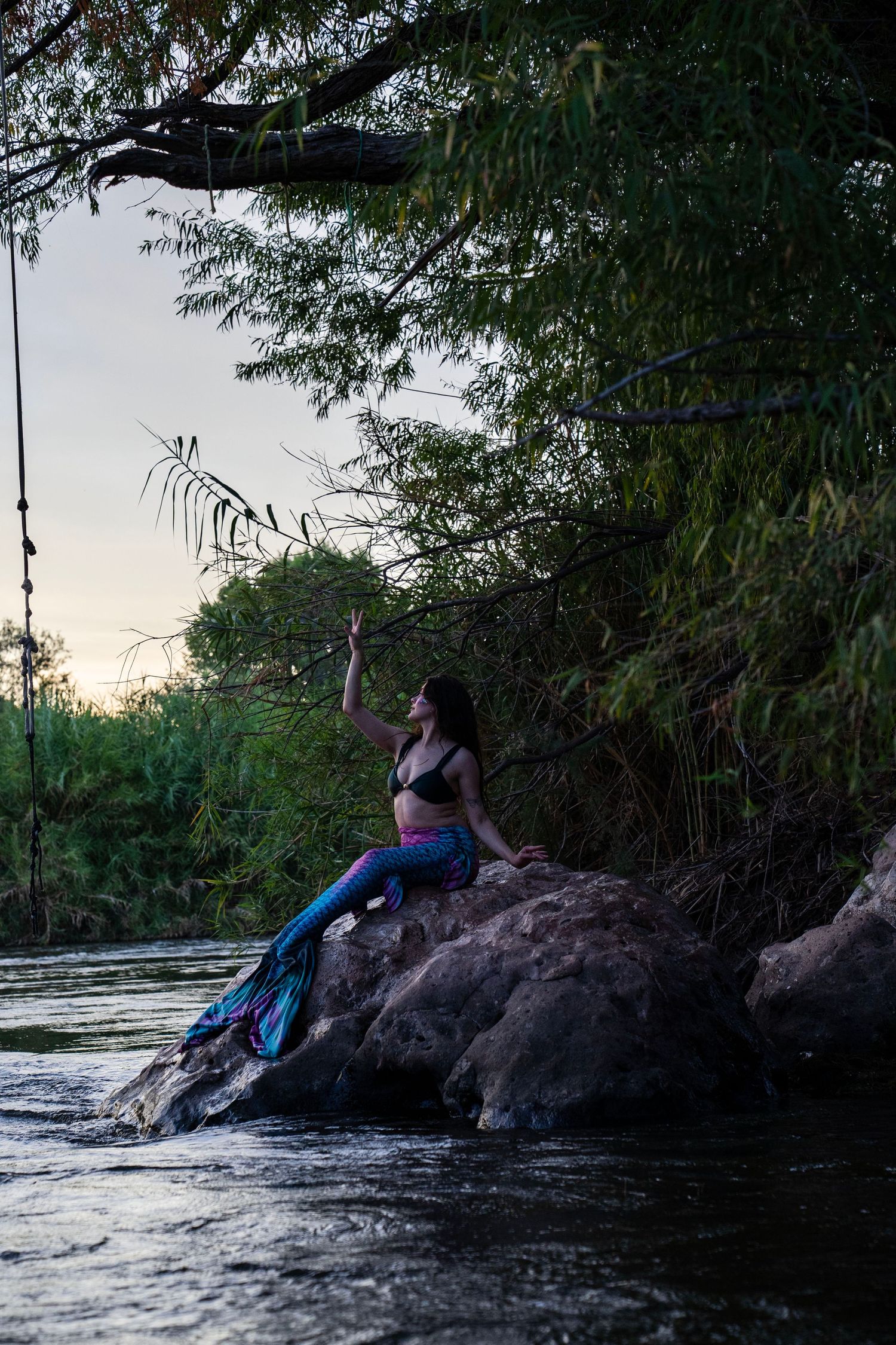 A woman dressed as a mermaid sits on riverside rocks at sunset, raising her hand toward lush green trees. Fantasy Photoshoot