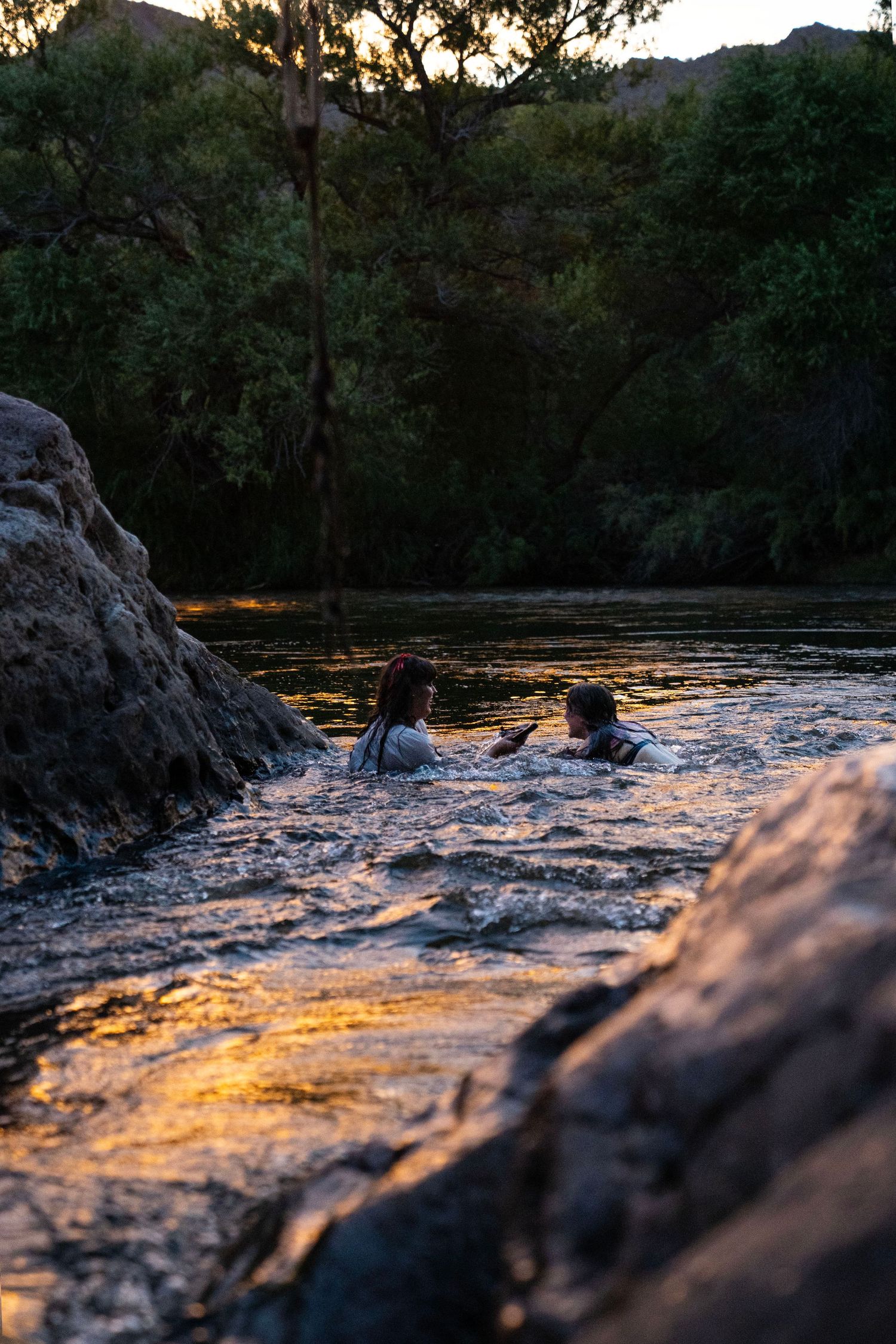 Two people swimming together in rocky river during colorful sunset. Fantasy photoshoot. Arizona photographer.