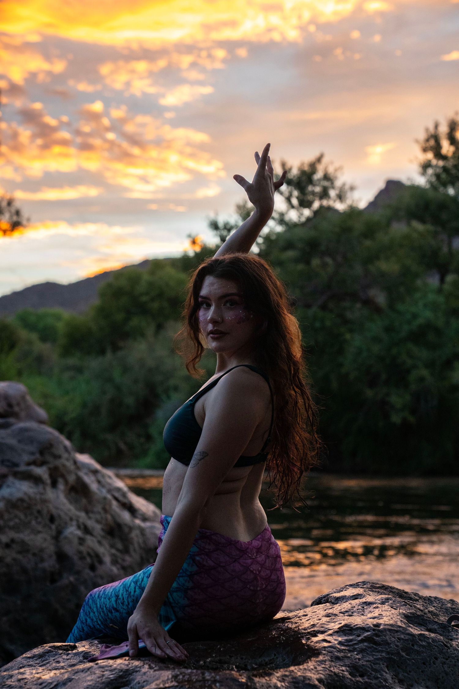 A woman in a mermaid tail costume poses on rocks with arm raised against a vibrant sunset sky. Mermaid photoshoot, fantasy photoshoot.