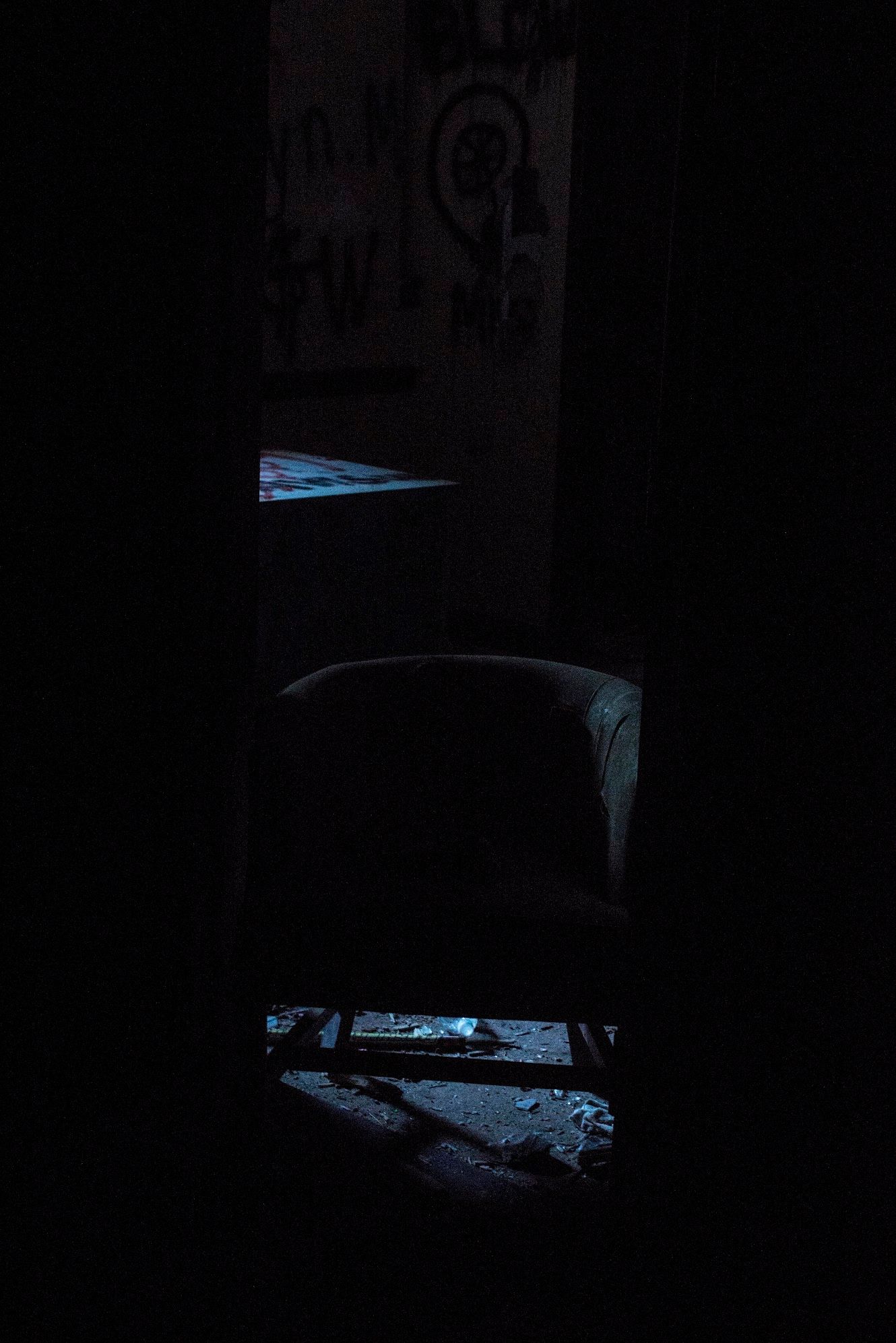 Dimly lit abandoned room with a worn armchair, debris-covered floor, and graffiti on the wall, seen through a narrow dark opening.