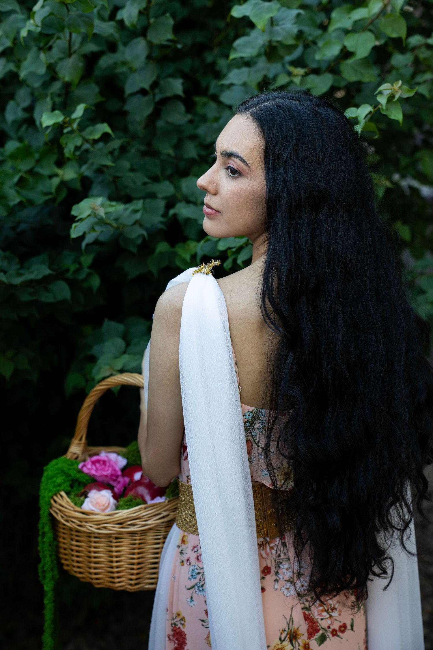 Woman with long wavy black hair in a floral peach dress and white sash holds a wicker basket of pink roses and greenery beside lush foliage.
