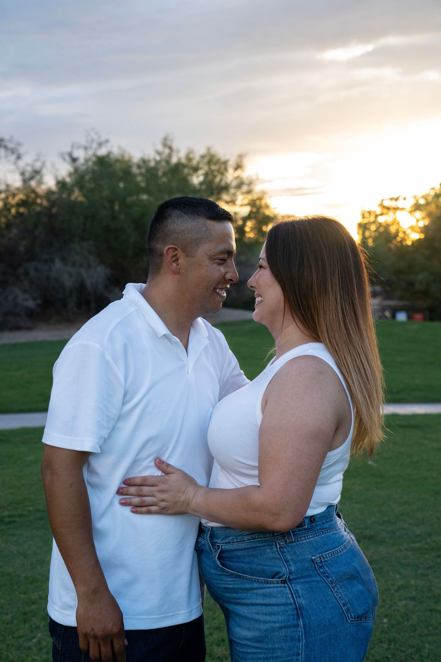 A smiling couple in white tops embrace at sunset in a lush green park, sharing a joyful moment together.