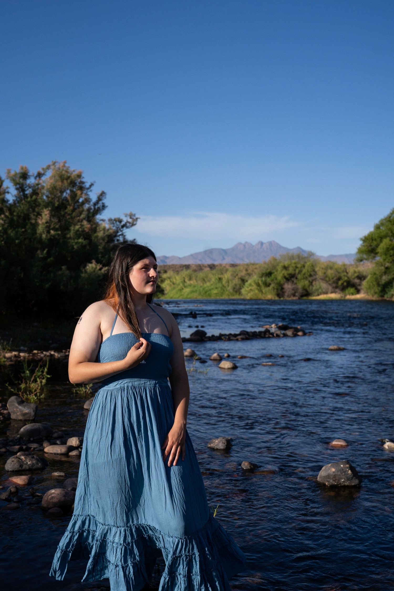 Woman in blue maxi dress standing by rocky river with mountain backdrop and clear blue sky.