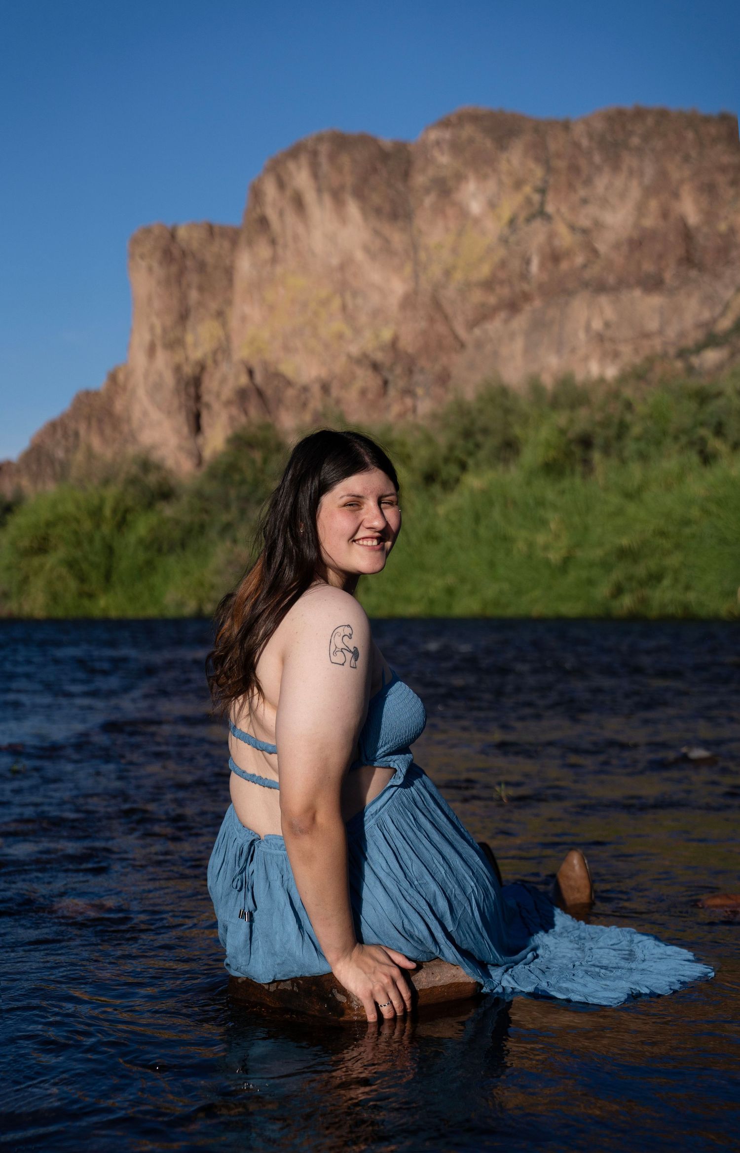 Woman in blue dress sitting in river water smiling, with rocky mountain cliff and green trees in background.