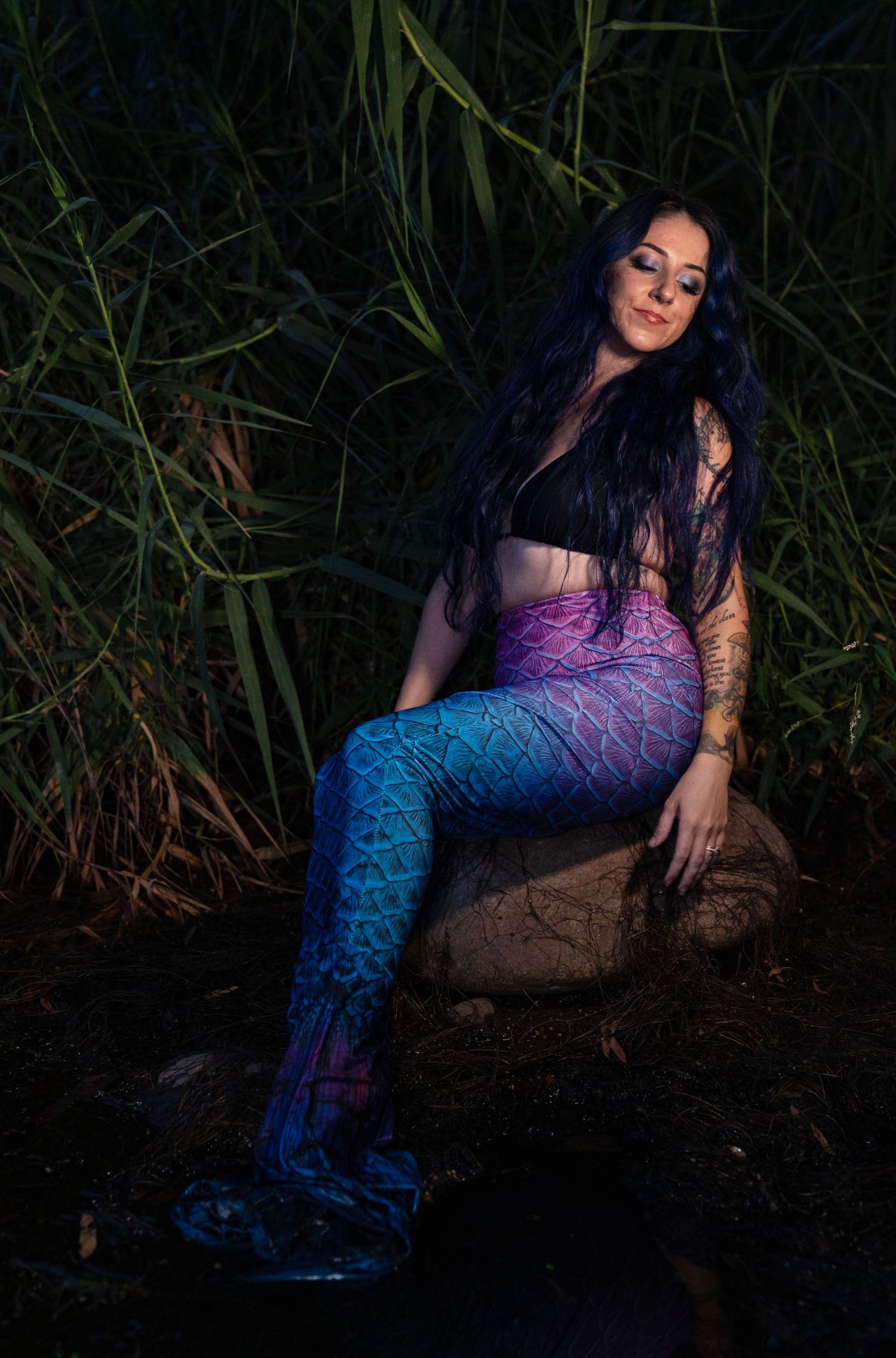 Woman in mermaid costume with blue-pink tail sits on rock among tall green reeds at night.