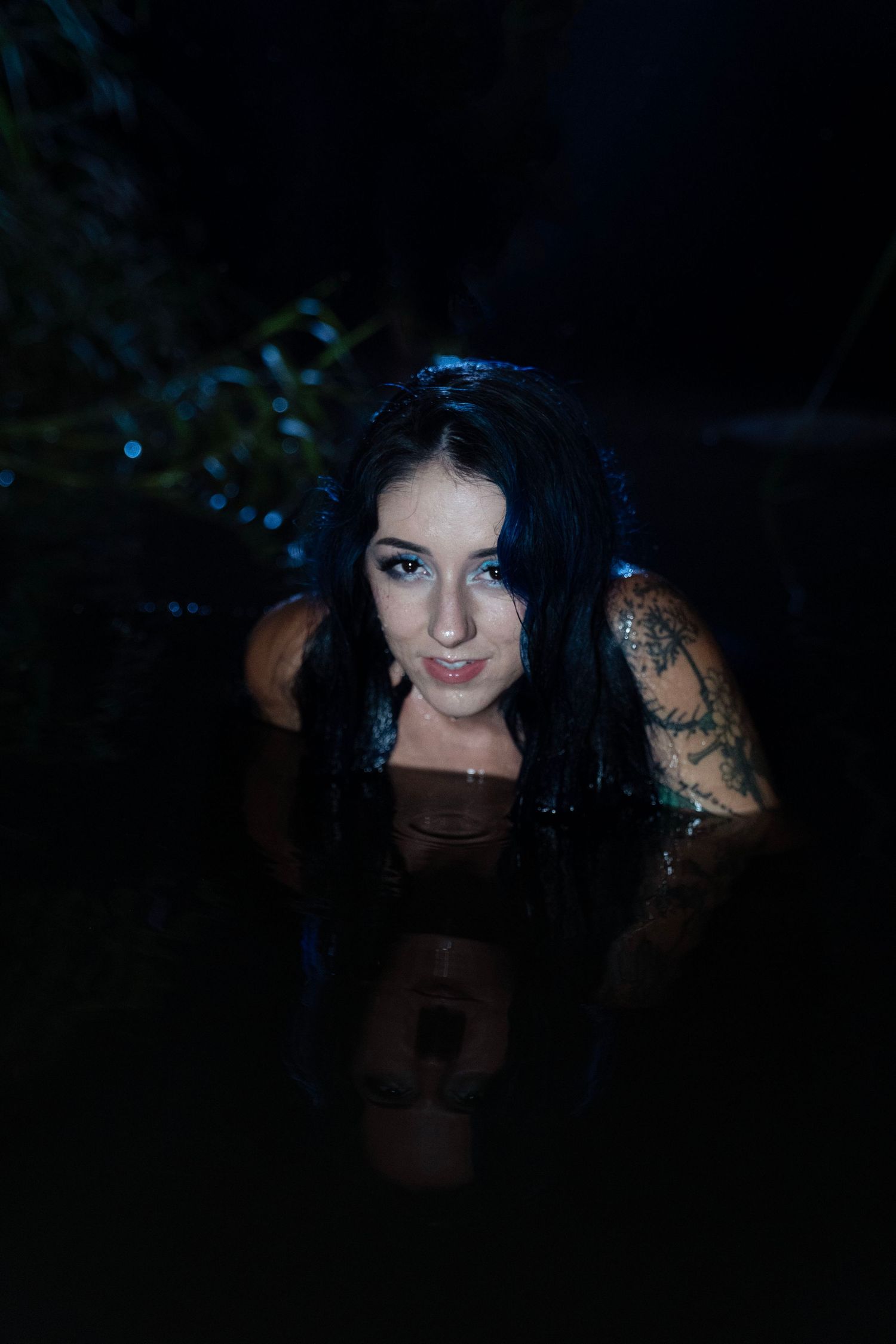 Tattooed woman with wet blue-black hair smiling while partially submerged in dark water at night.