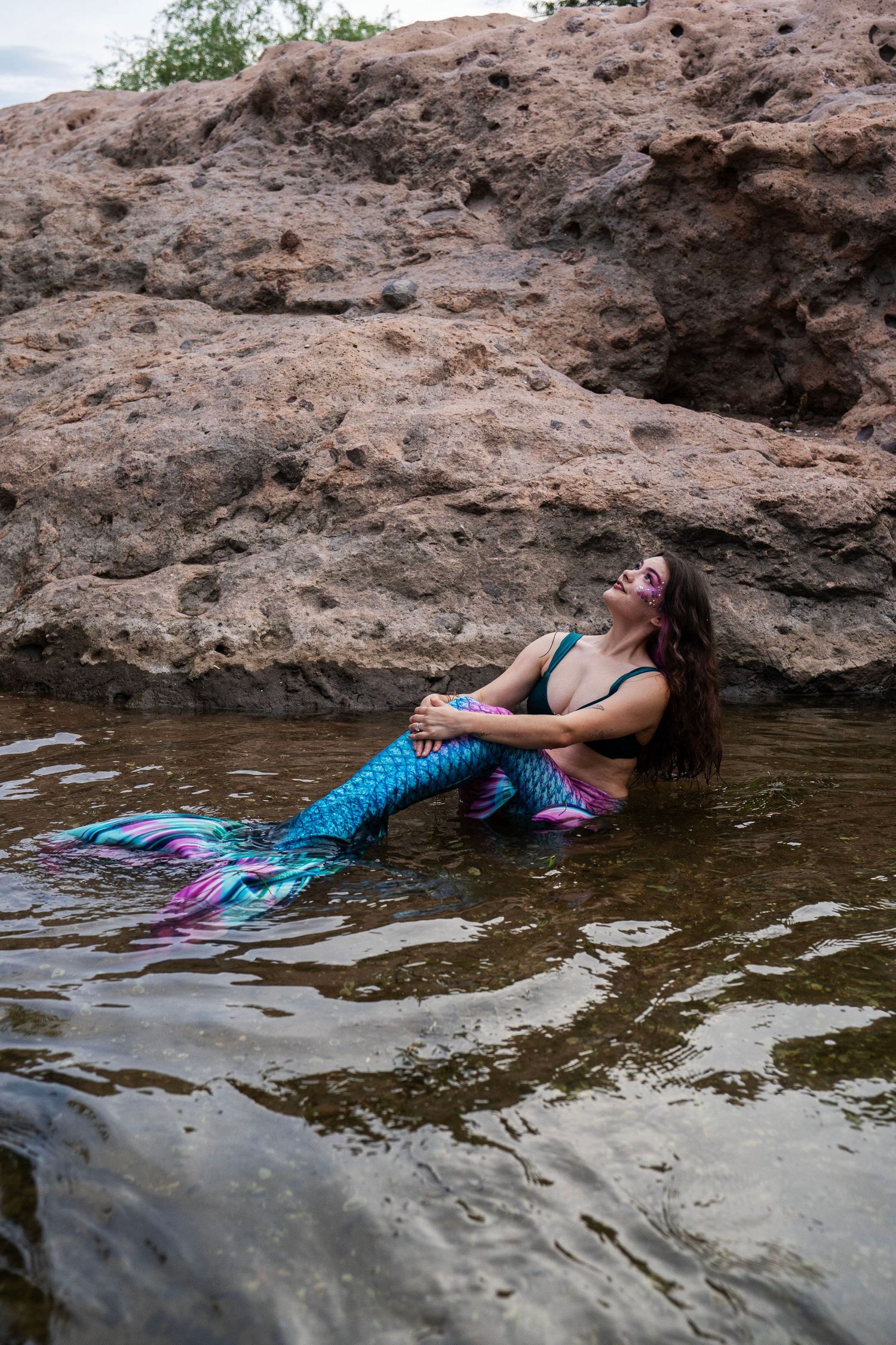A woman in a teal and pink mermaid tail costume reclines in shallow water against rocks. Fantasy Photoshoot