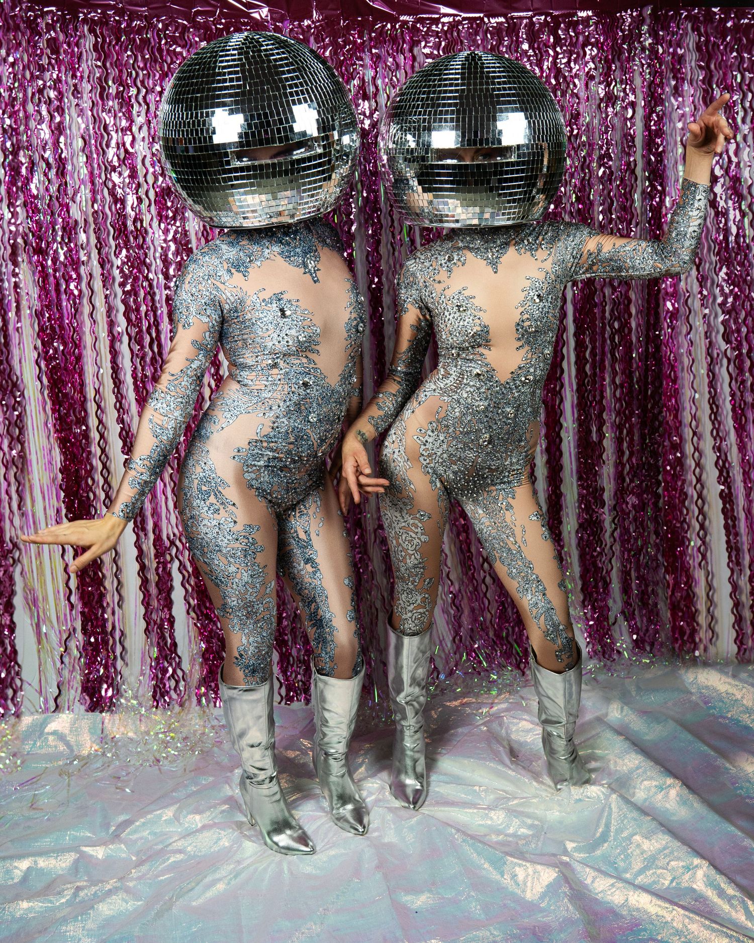 Two models in matching silver crystal catsuits and disco ball helmets pose together on pink backdrop.
