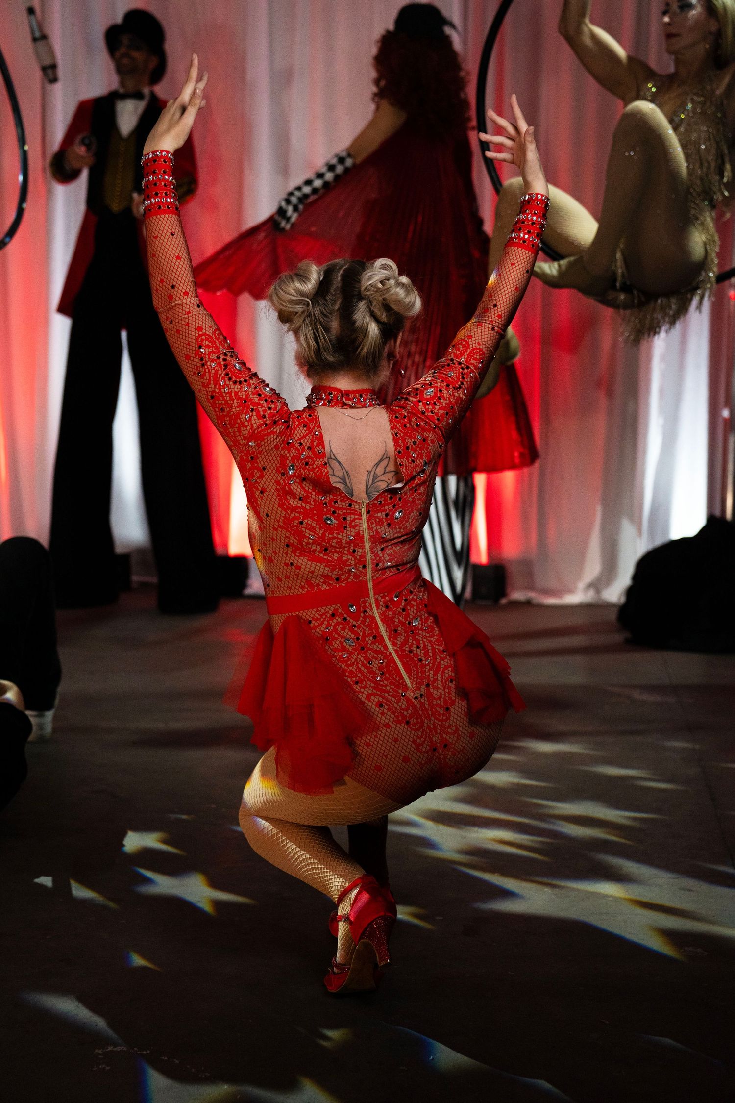 A dancer in a red sequined costume performs on a circus stage with aerial hoop artists in background.