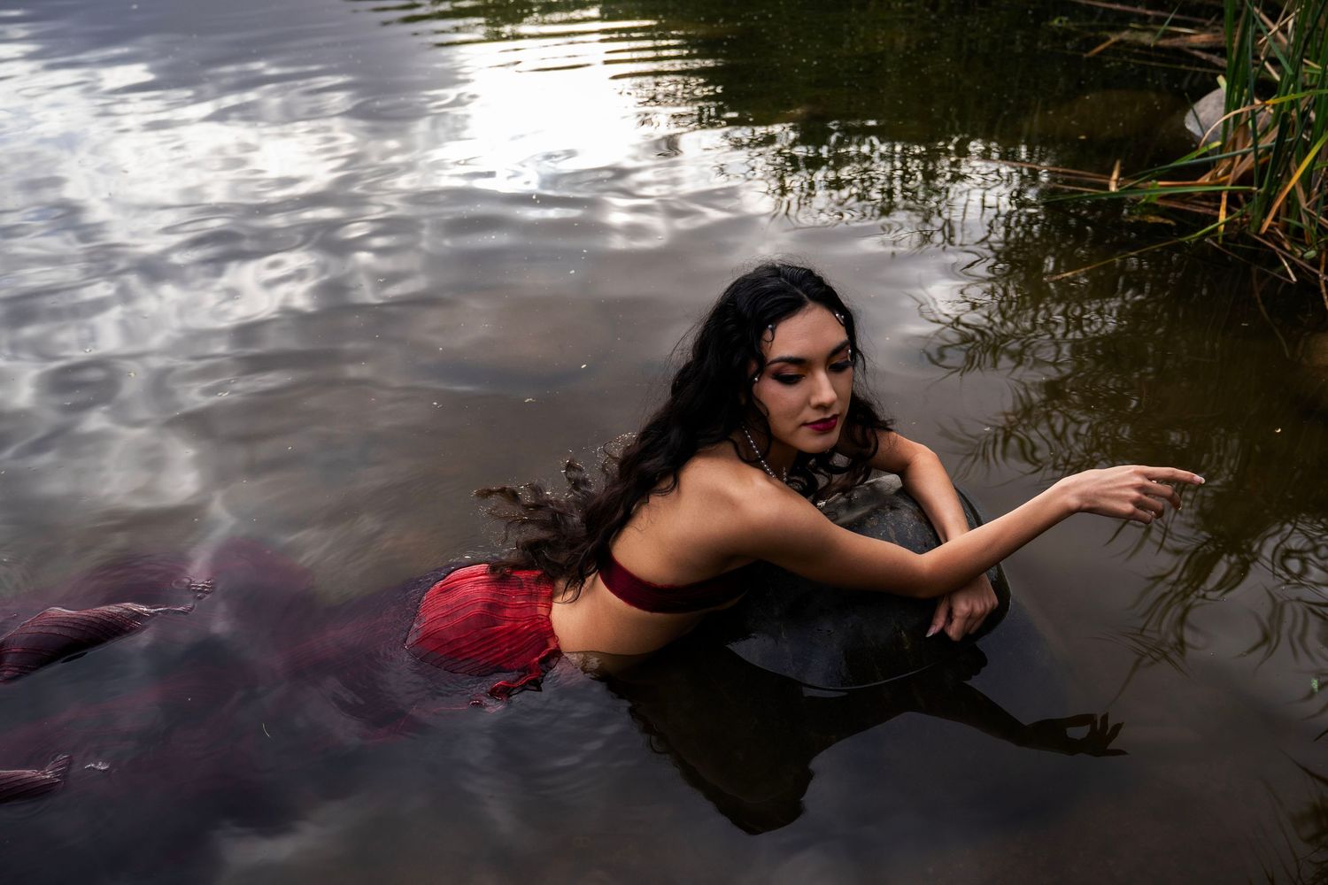 A woman in a red mermaid costume poses gracefully on a rock in a calm river surrounded by lush green trees.