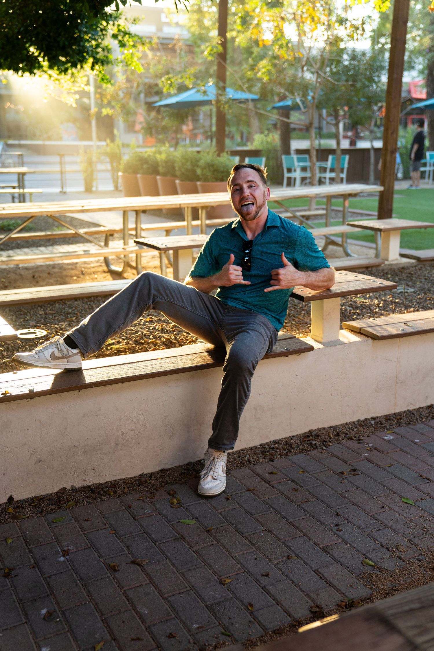 A cheerful man in a teal polo sits on a wall outdoors, giving thumbs up in a sunny park setting.