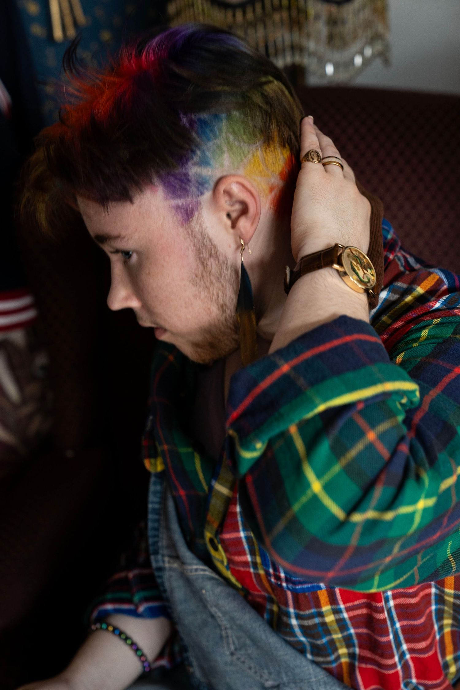 Person with rainbow tie-dye shaved sides hairstyle wearing plaid shirt and gold watch, shown in profile view.