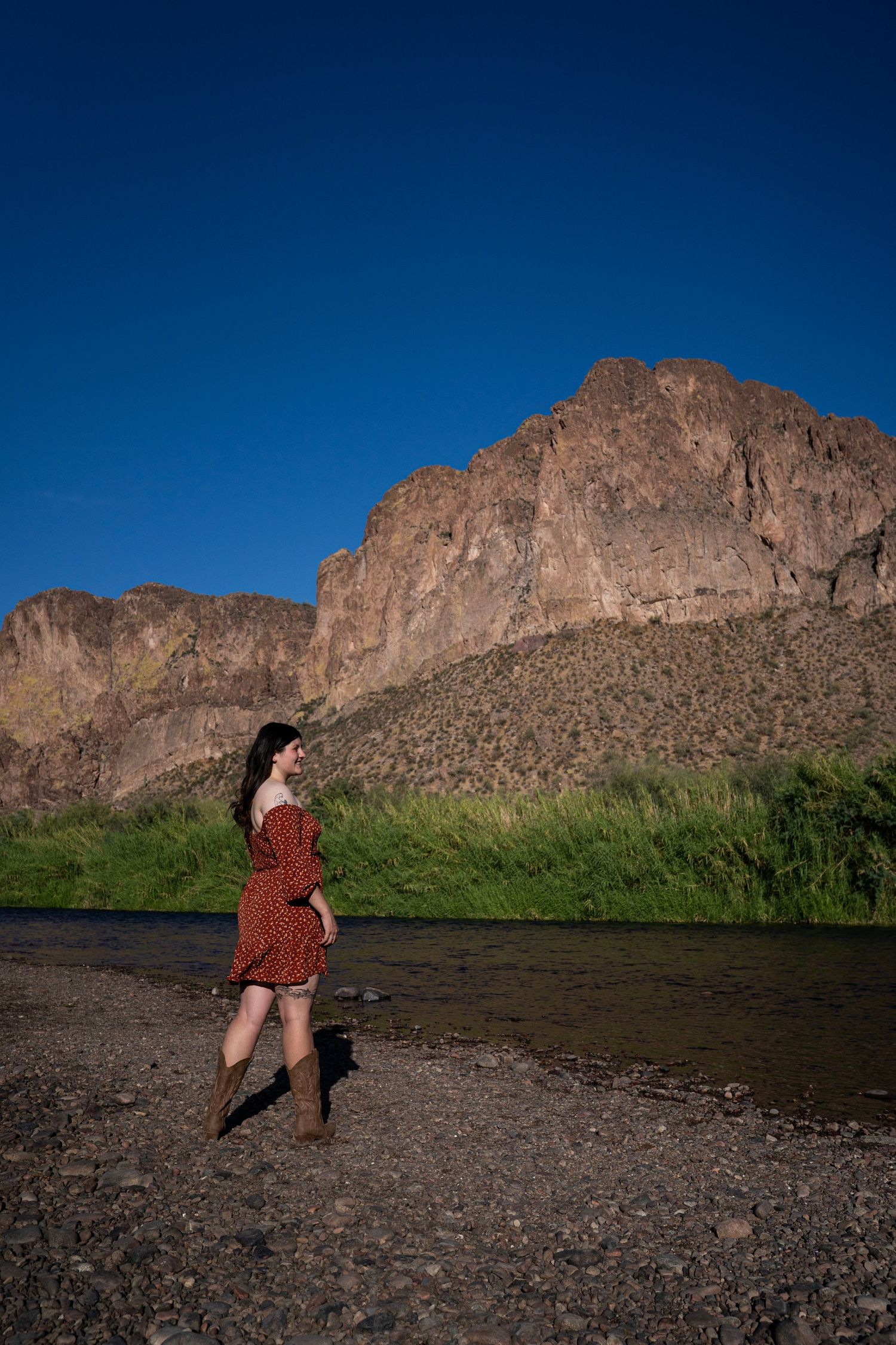 Woman in red dress standing by river with dramatic rocky mountain backdrop under clear blue sky.