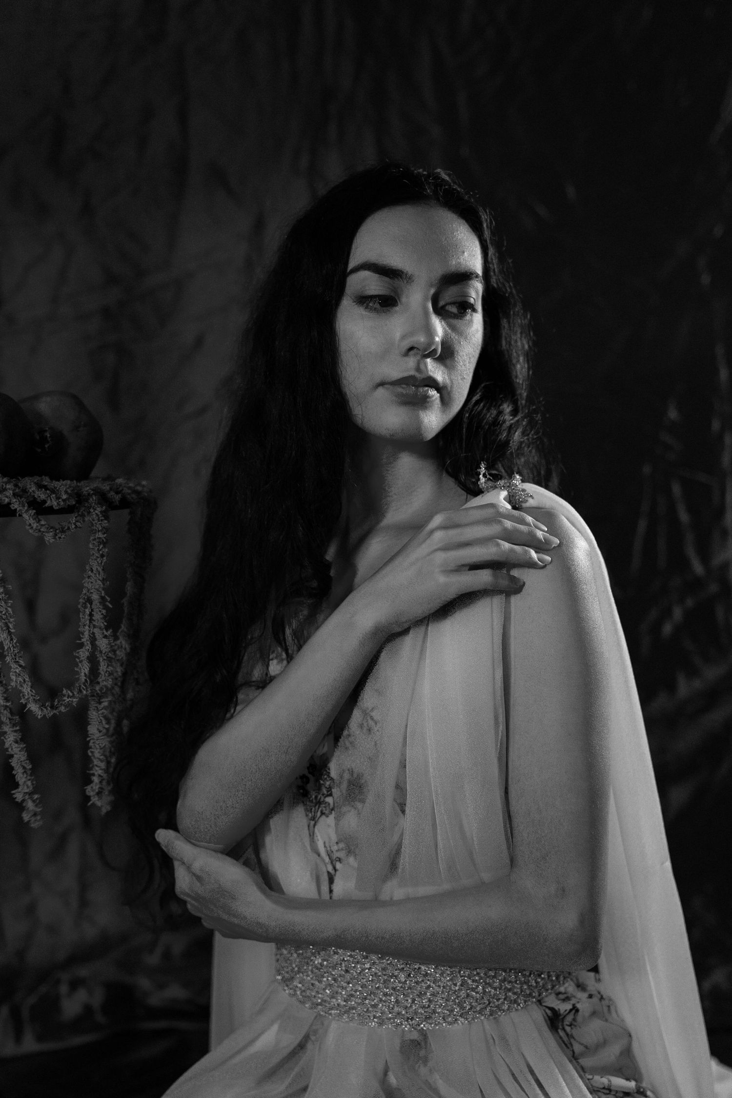 Black and white portrait of a woman with long dark hair in a flowing gown with beaded belt, arms crossed over her shoulder, gazing aside.