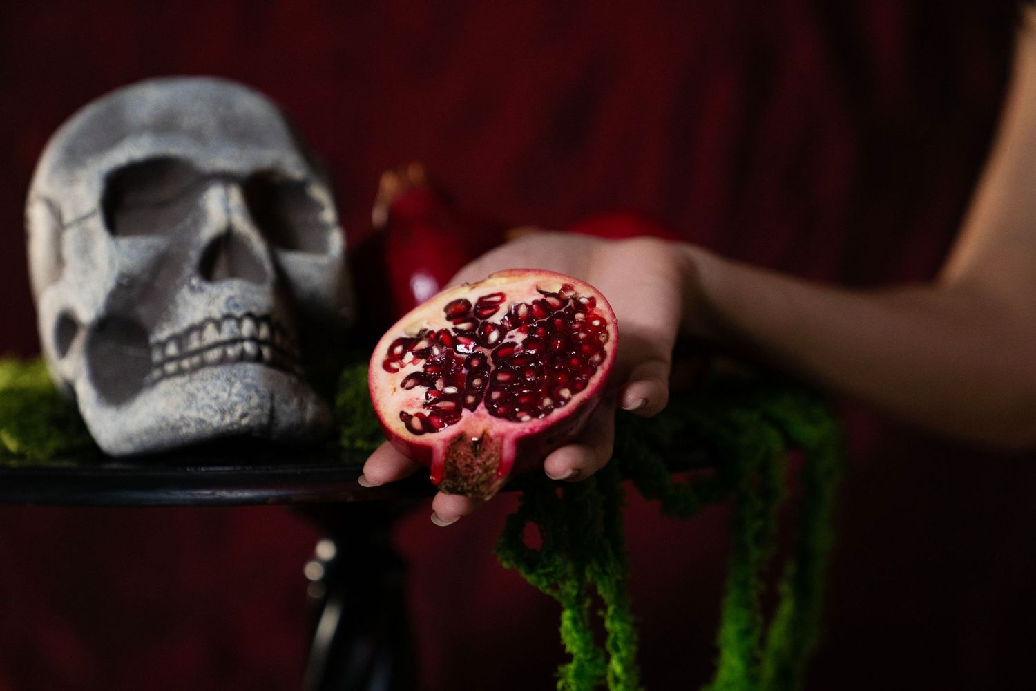 Hand holding a halved pomegranate with ruby red seeds beside a gray decorative skull and green garland against a dark red backdrop.