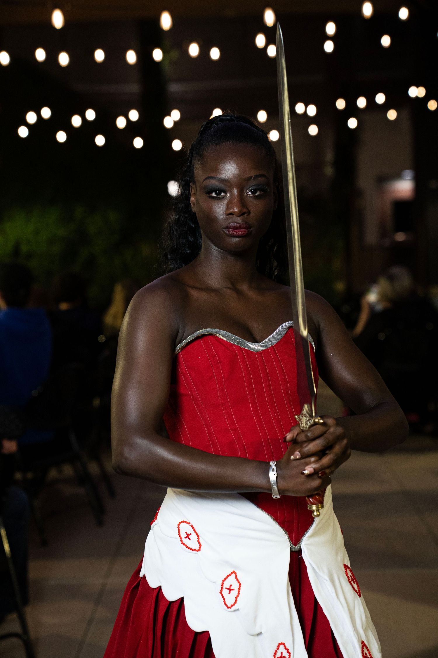 Woman in red and white gown holding a decorative sword under string lights at an outdoor evening event.