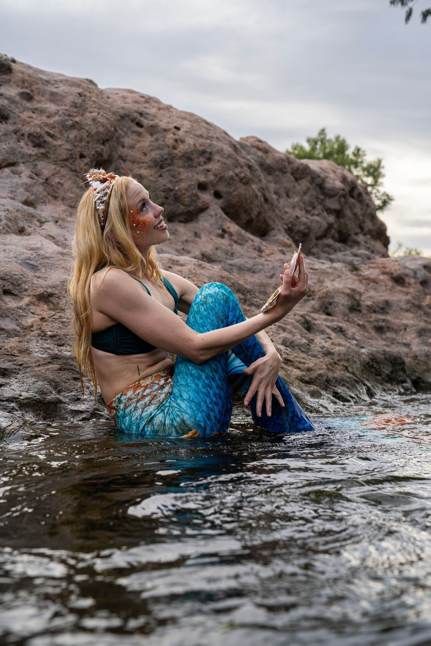 A blonde woman in blue mermaid costume sits in shallow water examining a seashell. Fantasy Photoshoot