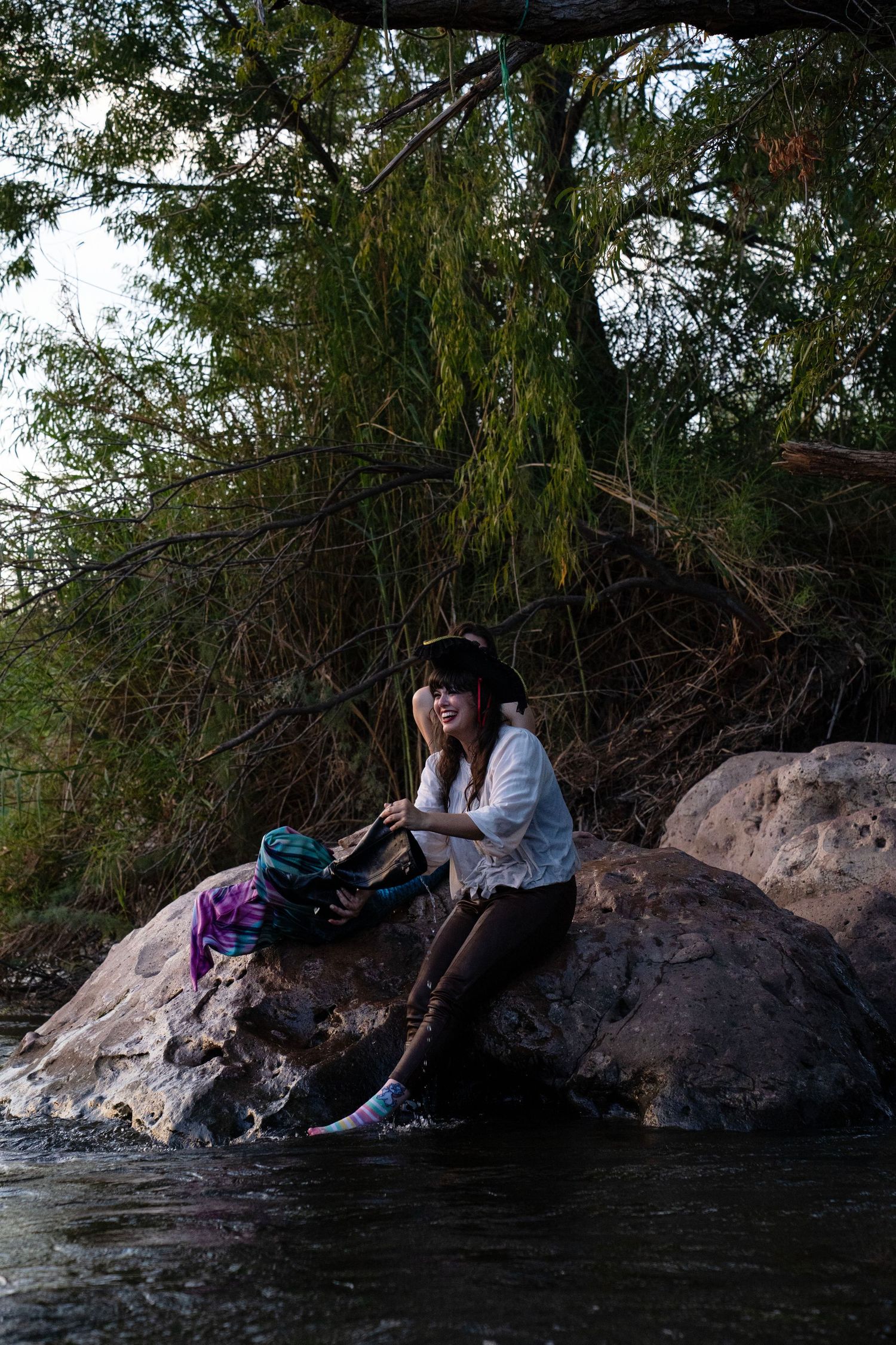 A woman in a pirate hat sits on rocks by a river, removing shoes near a colorful mermaid tail. Mermaid photoshoot, fantasy photoshoot.
