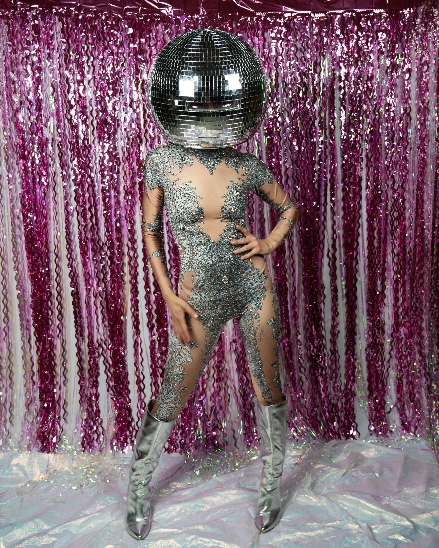Model in silver crystal bodysuit and boots poses with disco ball as head on pink backdrop.