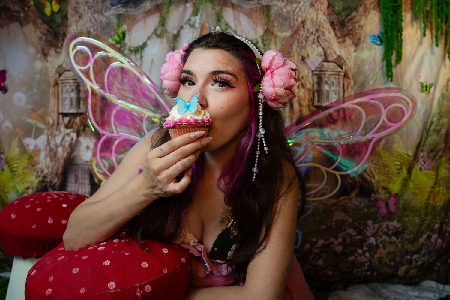 A fairy-costumed woman with colorful wings and pink flowers in her hair enjoys eating a cupcake in a magical setting. Fantasy Photoshoot