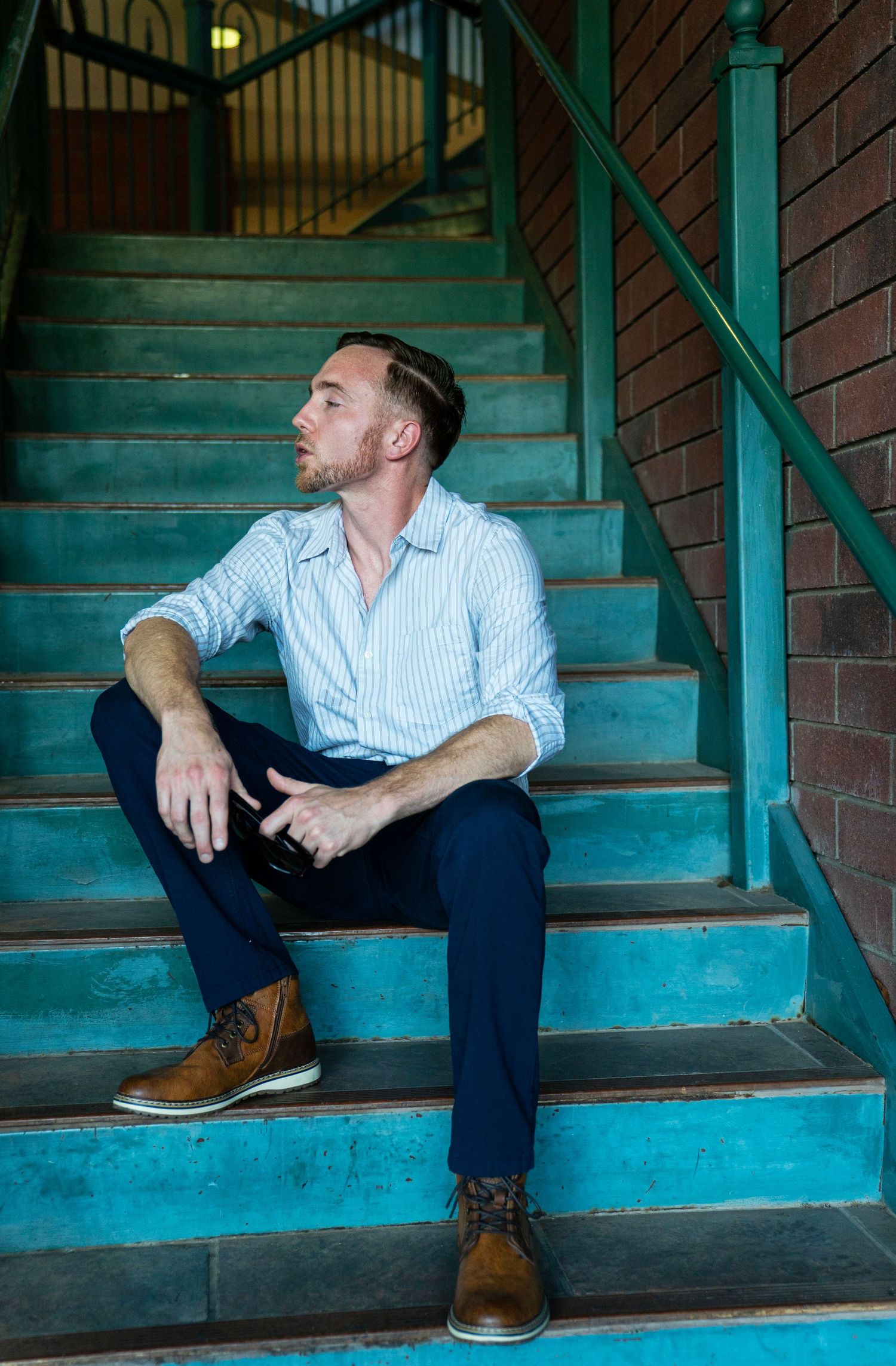 Man in striped shirt and navy pants sits on teal stairs, gazing upward thoughtfully, wearing brown leather boots.