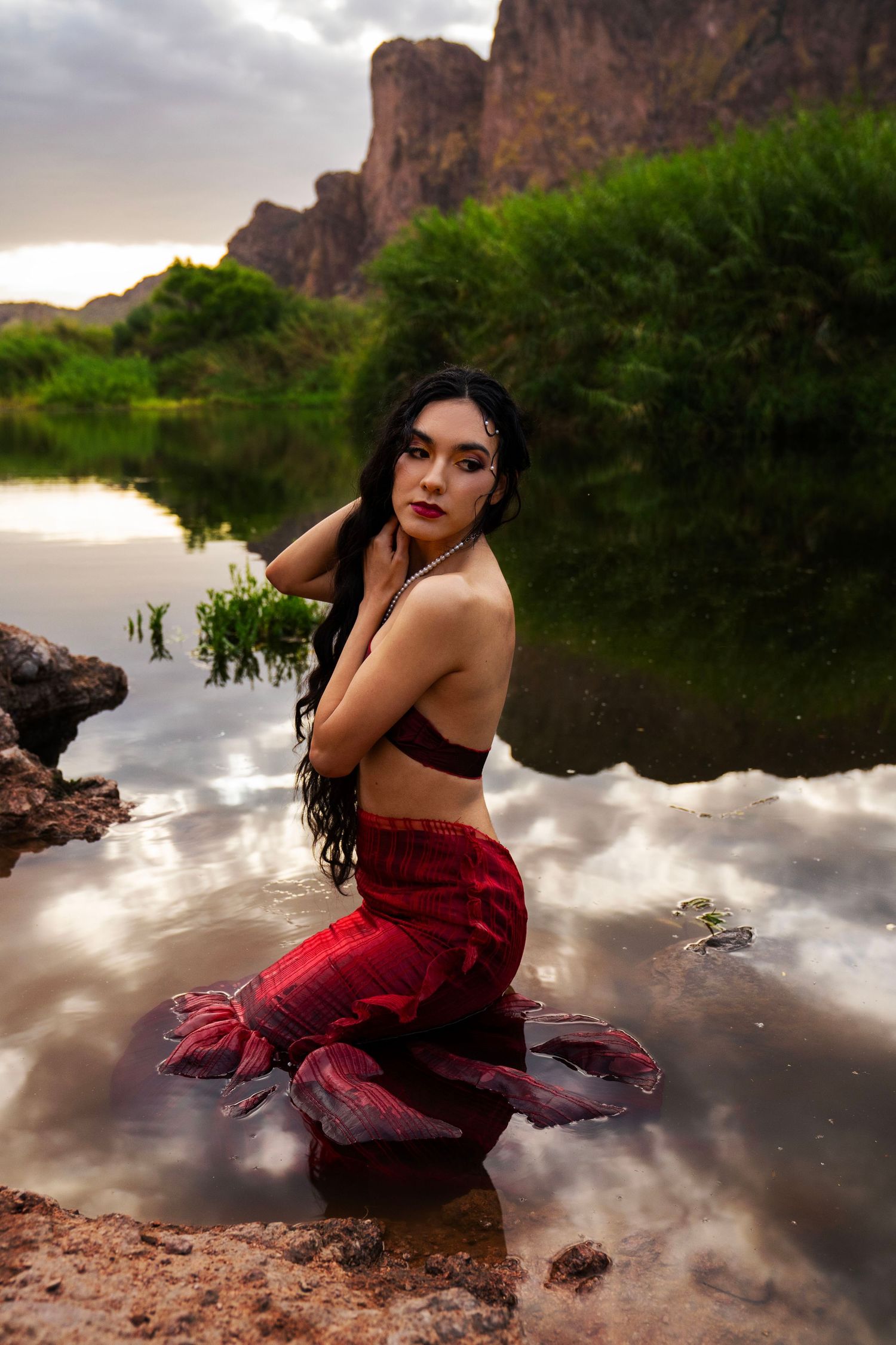 A woman with black hair and red lips sits in shallow water wearing a red skirt with rocky cliffs and greenery behind her.