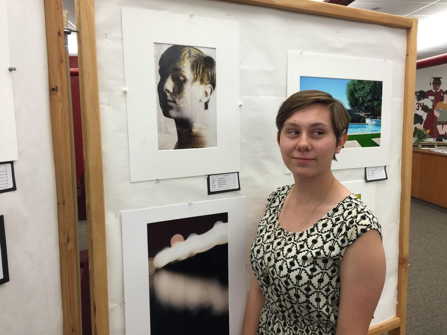 Young woman with short hair in a patterned sleeveless dress stands beside framed black-and-white photographs at an art exhibit.