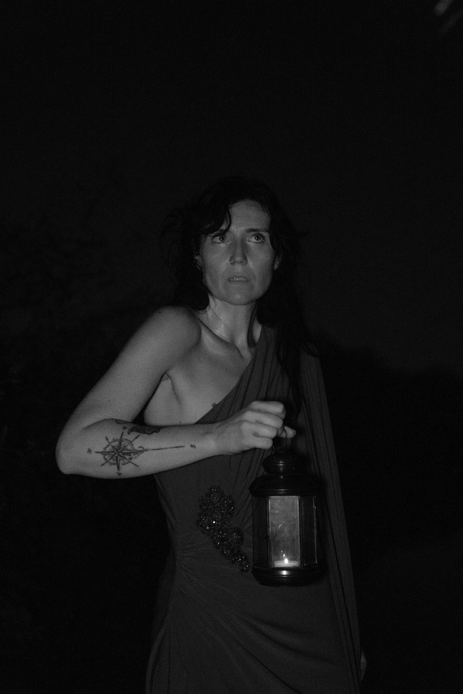 Black and white portrait of a woman in a draped one-shoulder dress holding a lantern, showing a compass tattoo on her forearm.
