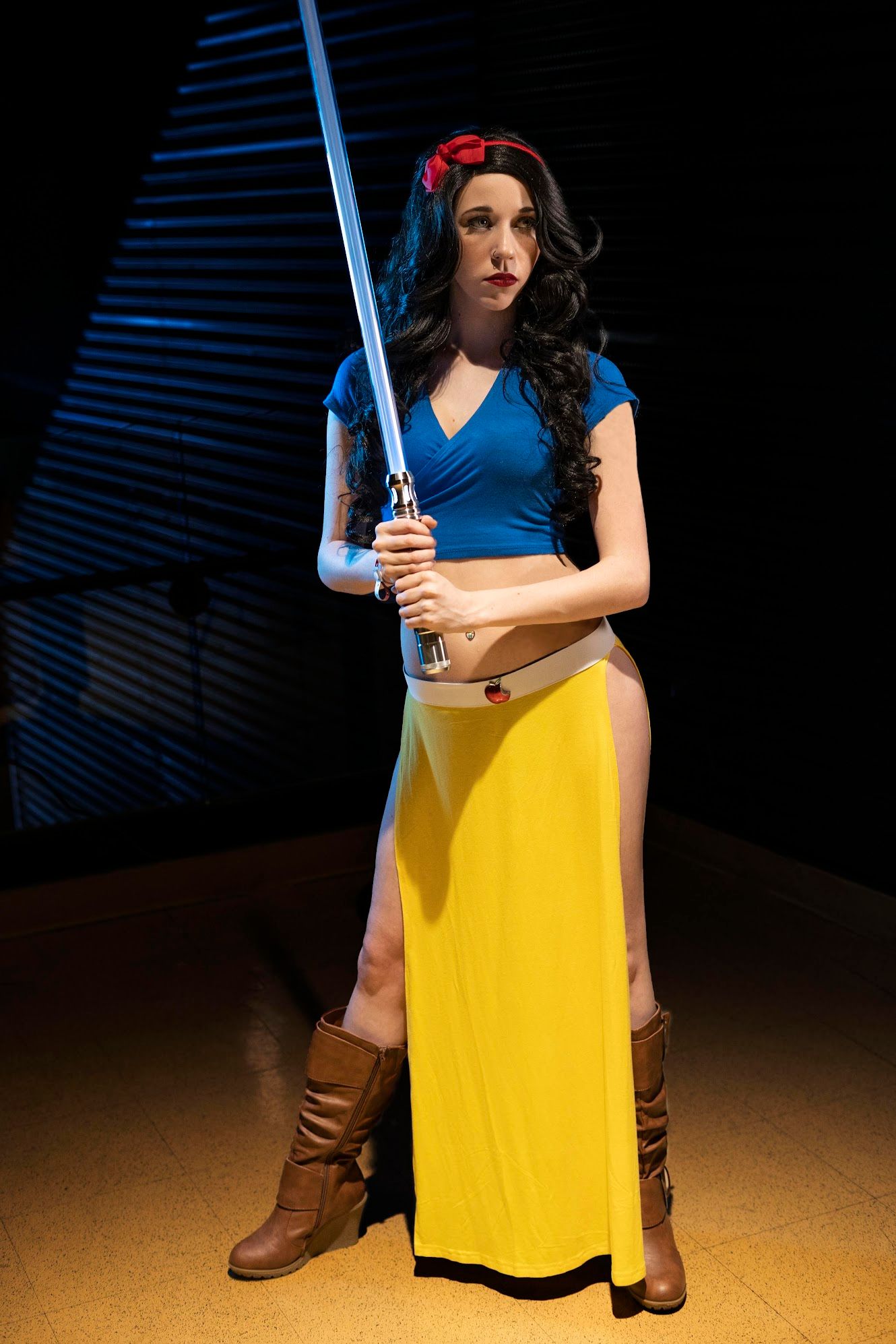 Cosplayer in a Snow White-inspired Jedi costume with red hair bow, blue top, yellow skirt and brown boots, holding a blue lightsaber.