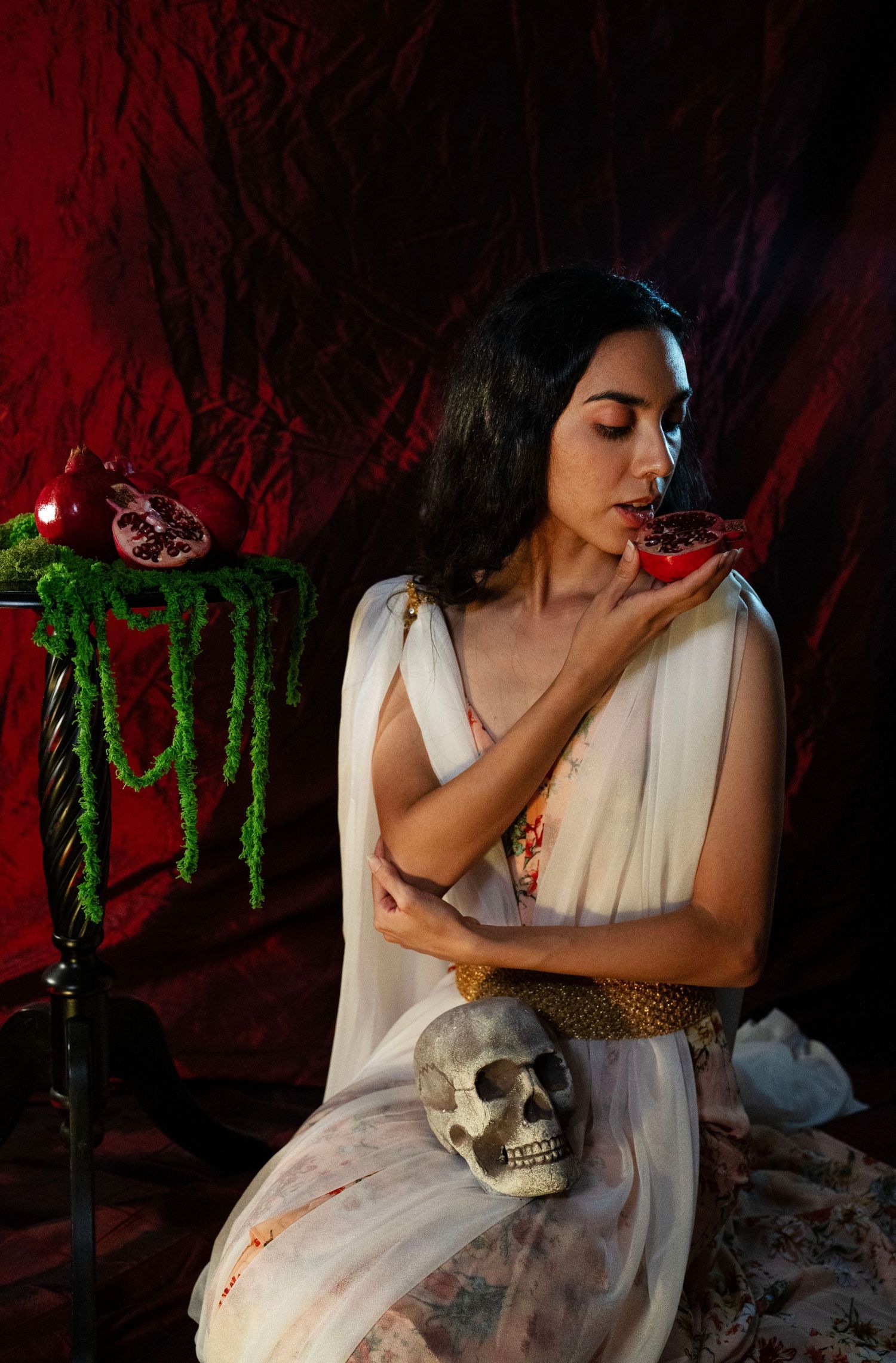 Woman in a flowing white gown holds a split pomegranate to her lips, a skull in her lap, against a dark red velvet backdrop.