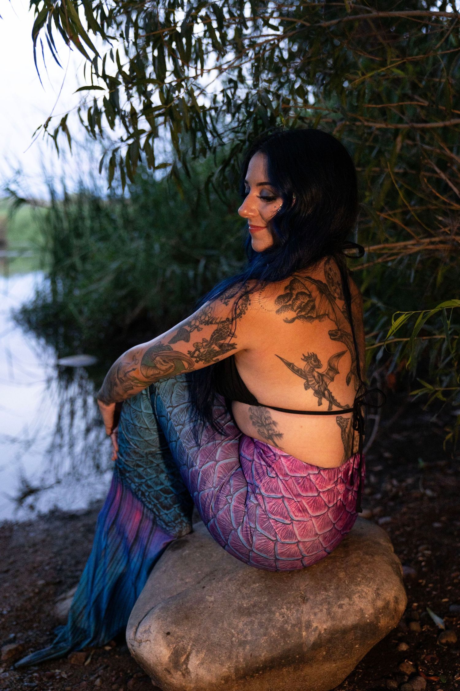 Tattooed woman in mermaid costume sits on rock by water at dusk.