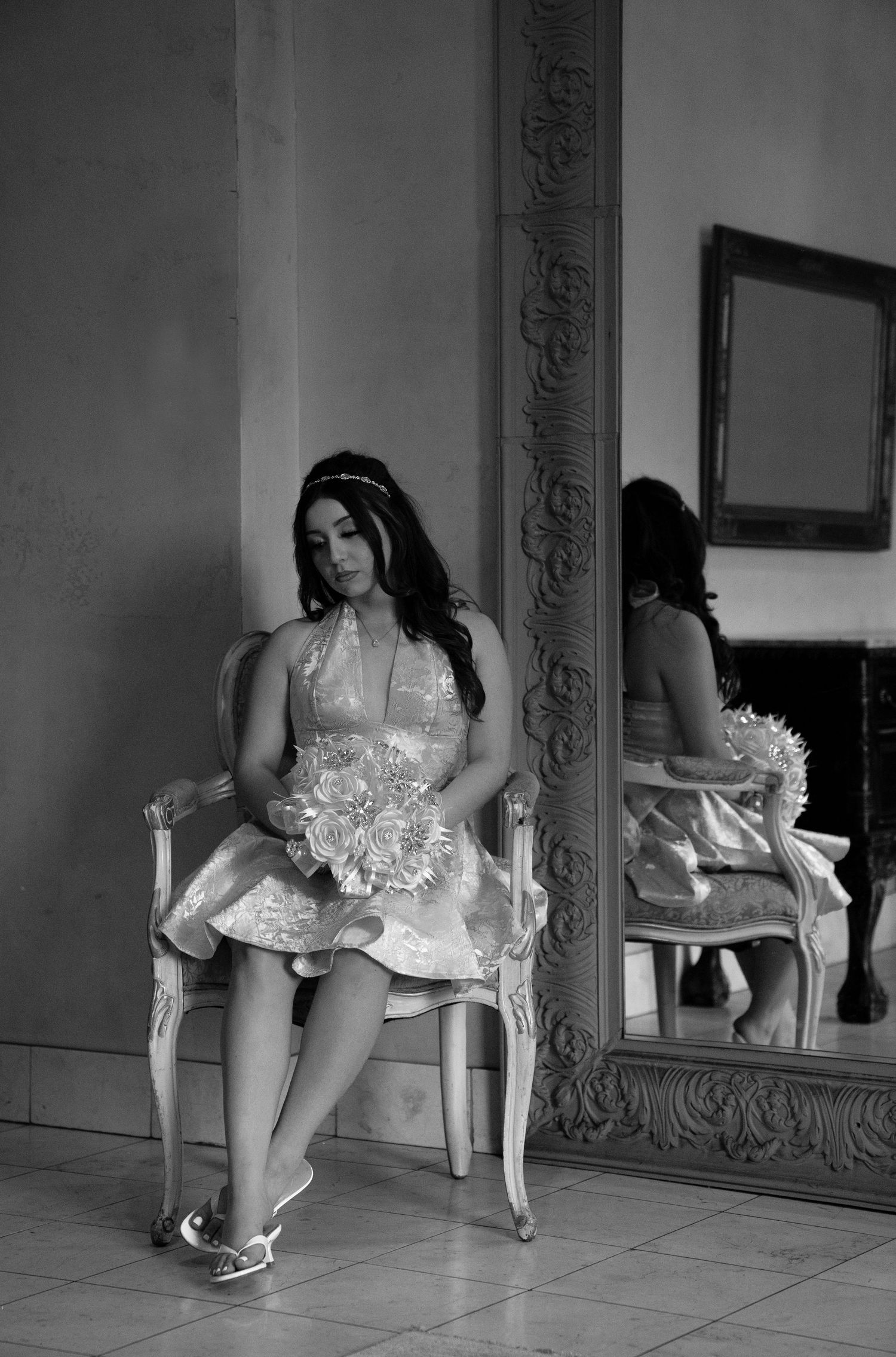 Young woman in formal dress holding bouquet seated in ornate chair beside large mirror, black and white photo.