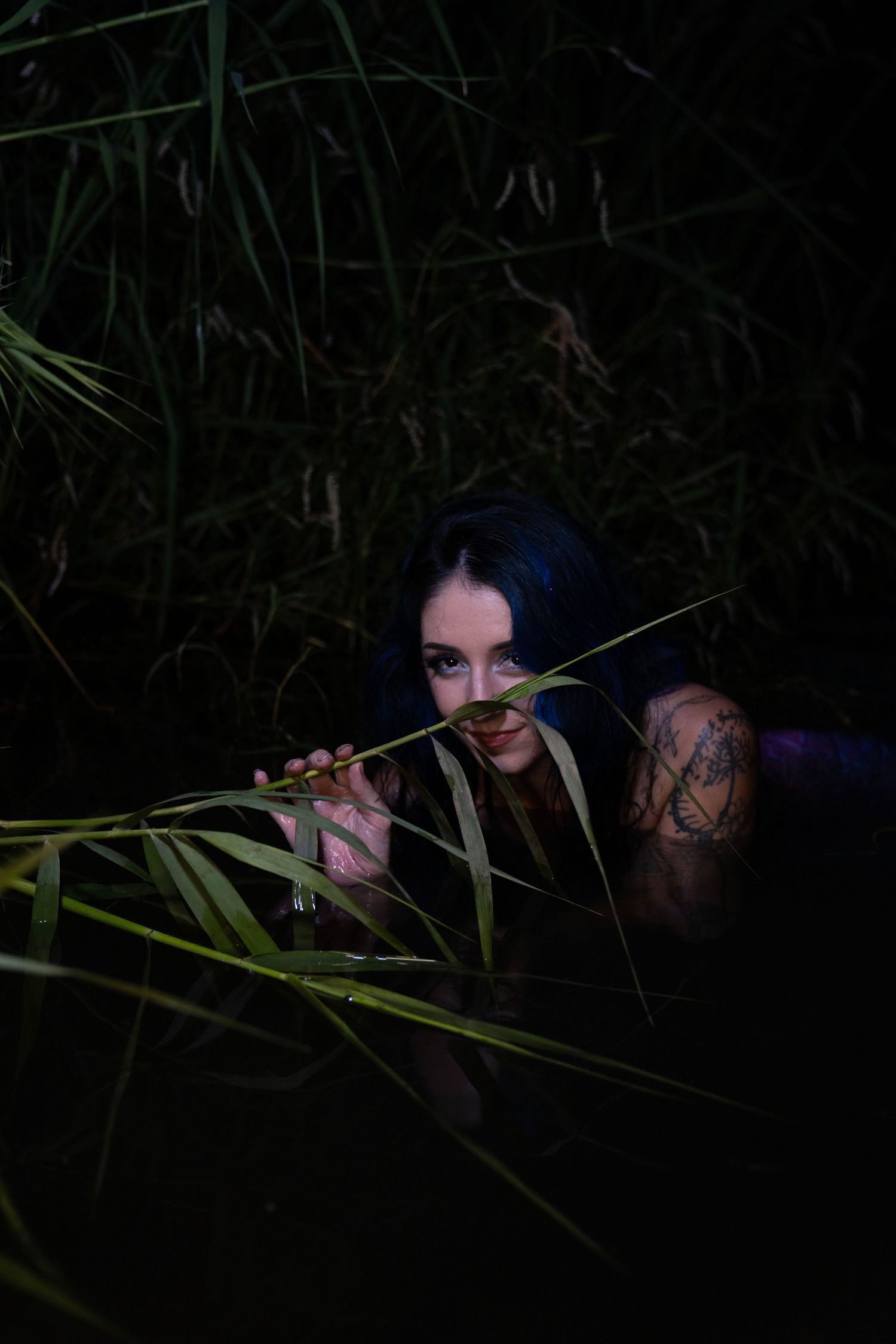 Woman with blue hair and tattoos peers through tall green reeds at night in a mysterious outdoor setting.