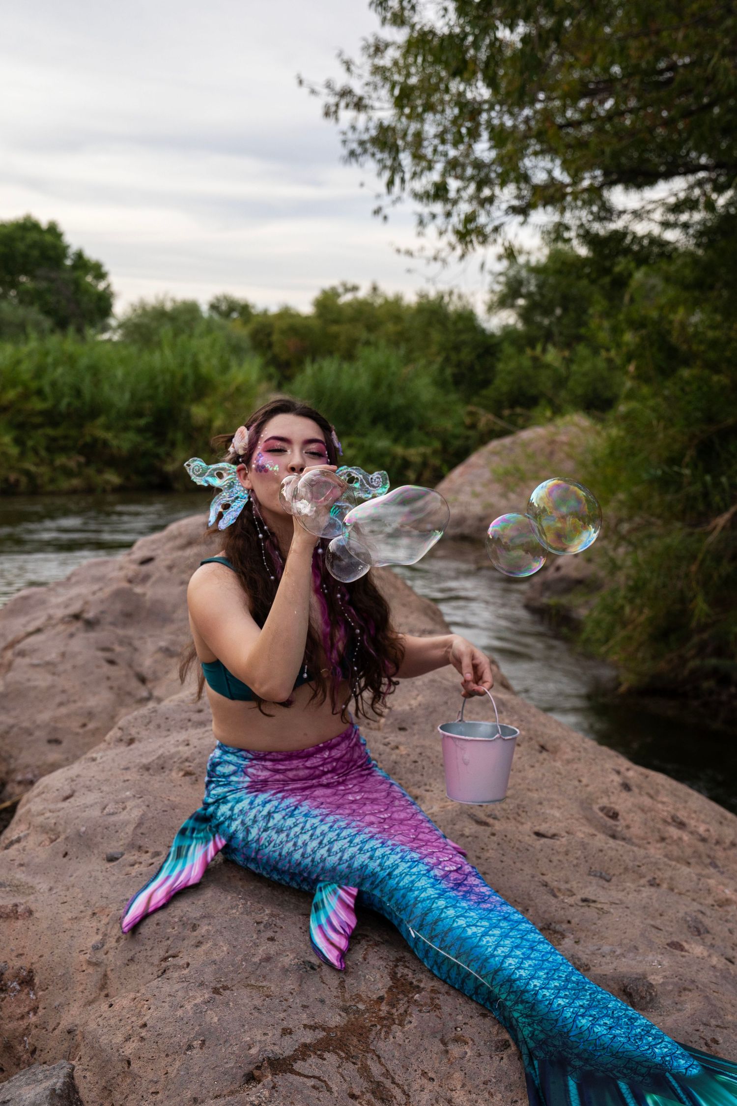 Woman dressed as a mermaid with colorful tail blowing soap bubbles while sitting on rocks near a river. Fantasy Photoshoot