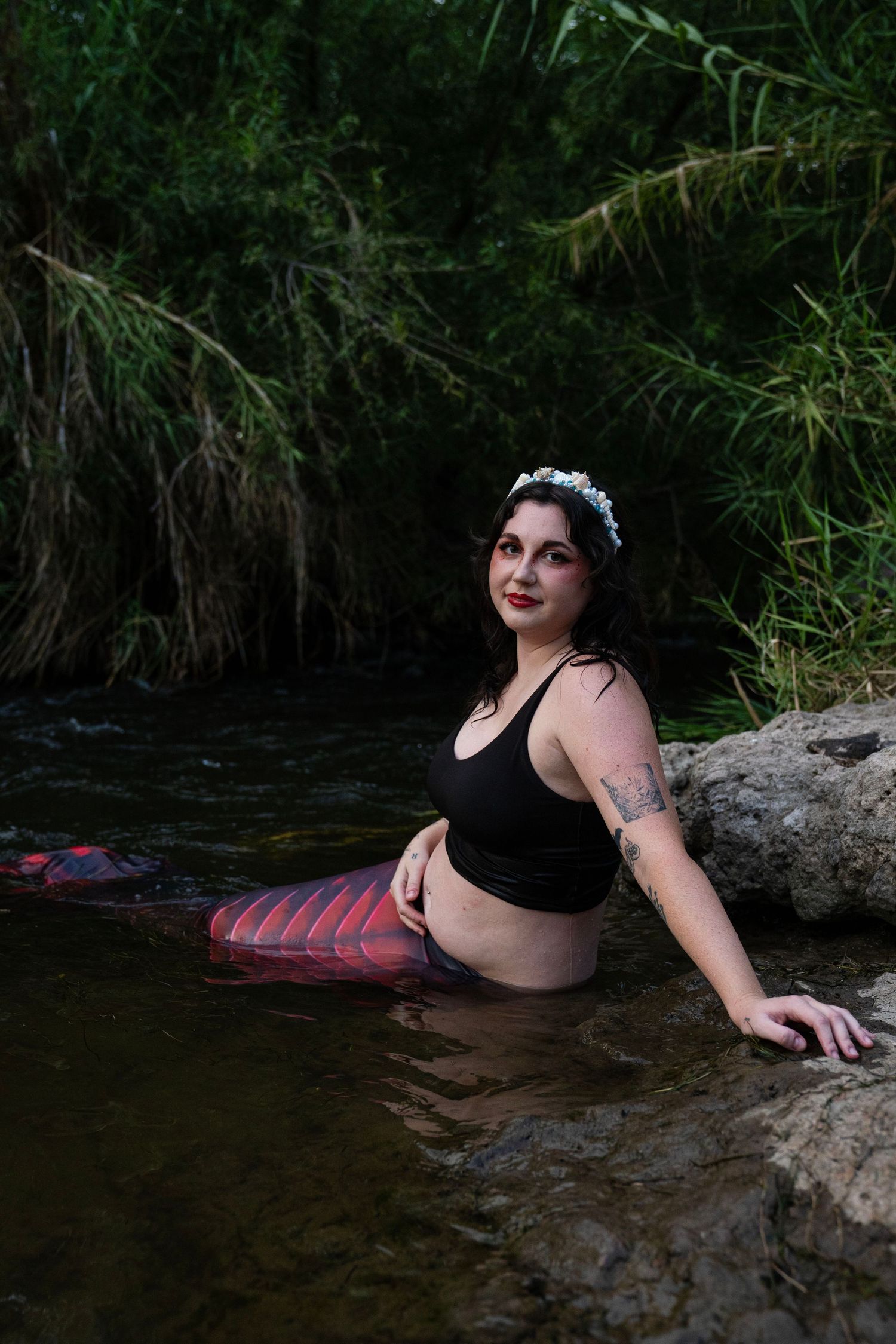 Tattooed woman in black top wearing white floral crown with red mermaid tail in water. Fantasy Photoshoot
