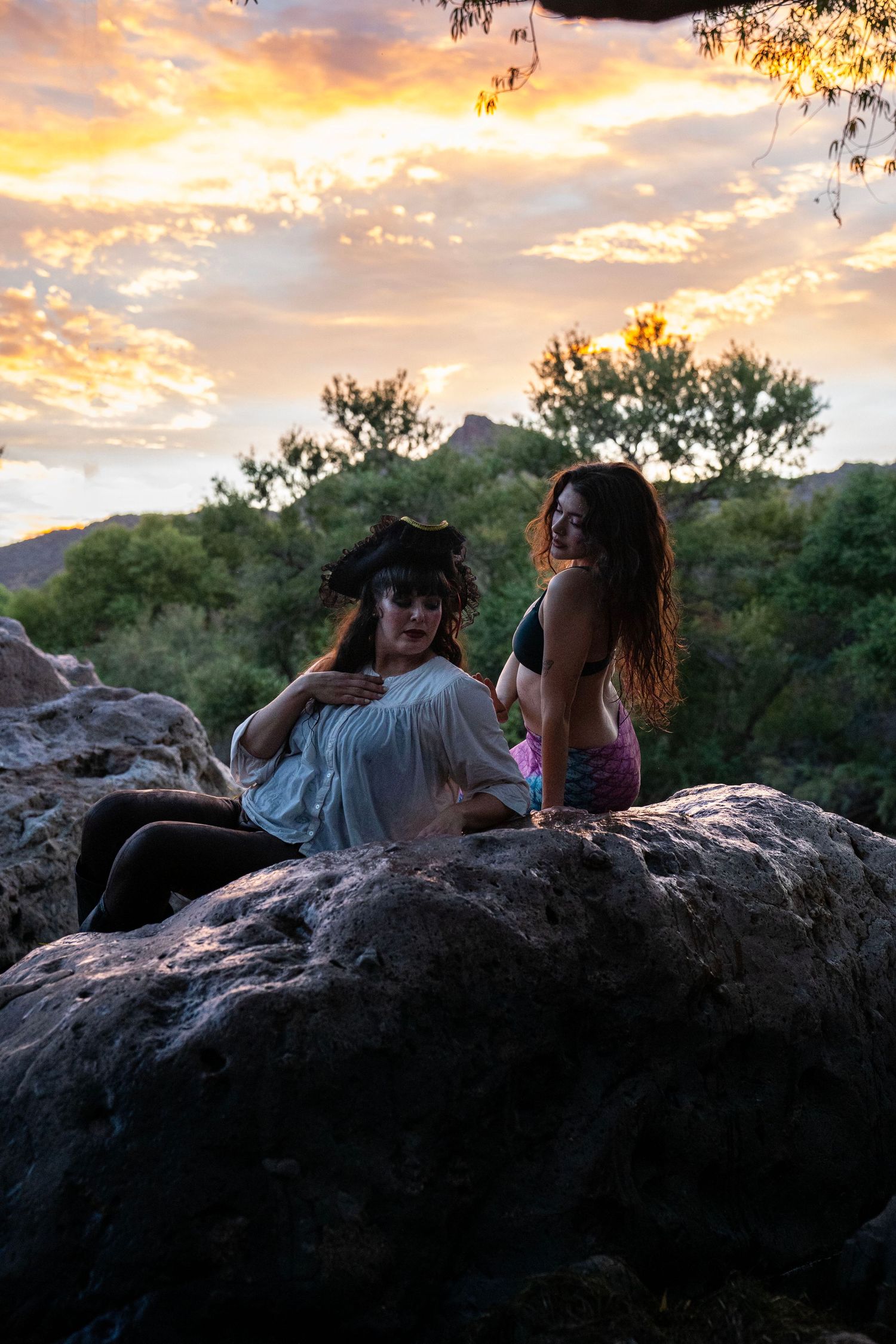 Two women in fantasy pirate and mermaid costumes sitting on rocks at golden sunset. Mermaid photoshoot, fantasy photoshoot.