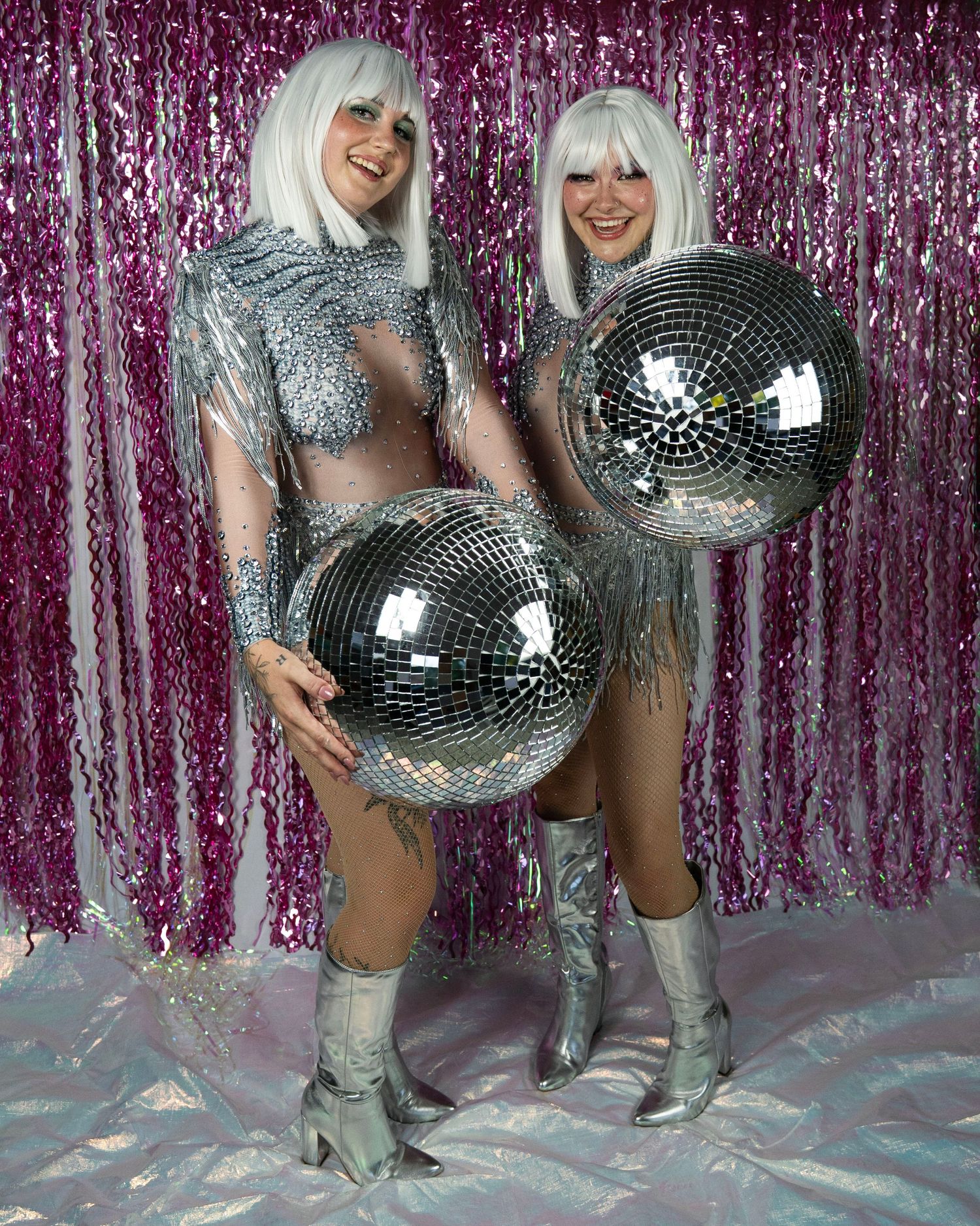 Two women in silver sequin disco costumes and white wigs stand holding large mirror balls against pink tinsel backdrop.