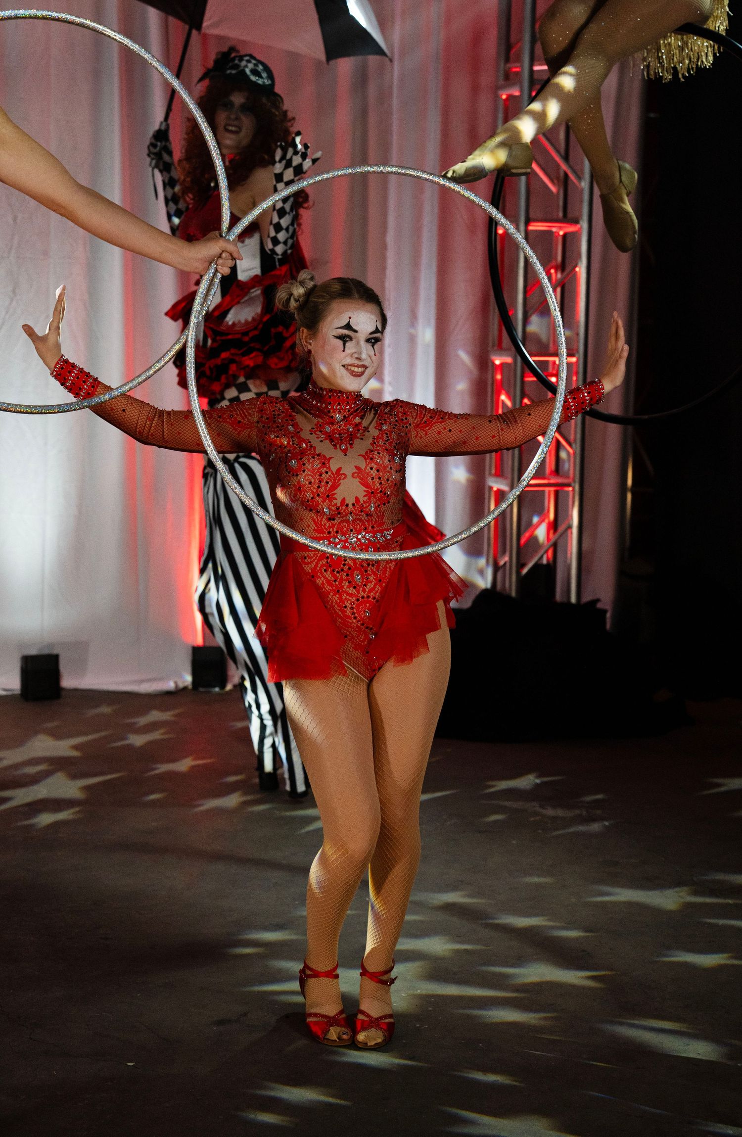 Female circus performer in red sequin costume spinning a silver hula hoop with mime-style makeup.