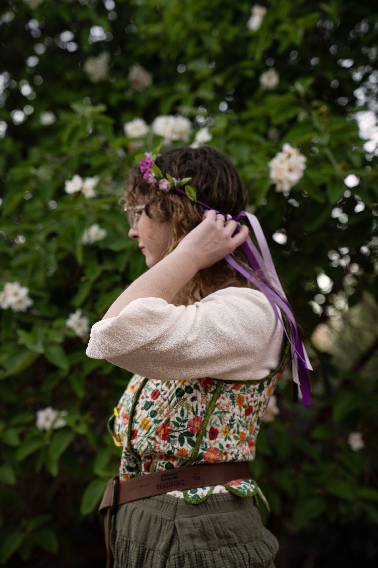 Woman in a cream blouse and floral vest adjusts a purple ribbon flower crown while standing before a blooming white-flowered shrub.