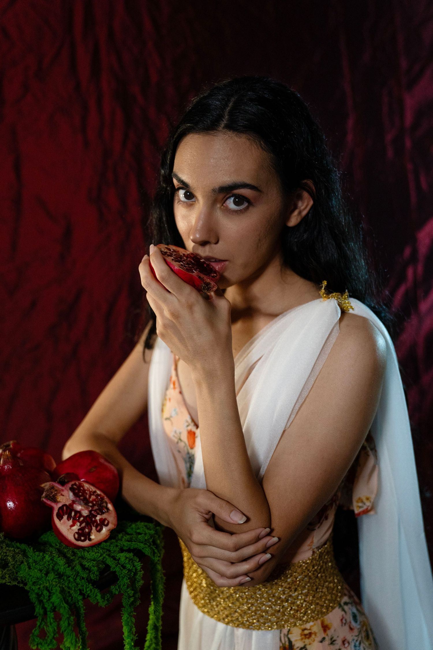 Woman in a white draped gown with gold belt holds a pomegranate half to her lips against a deep red velvet backdrop.