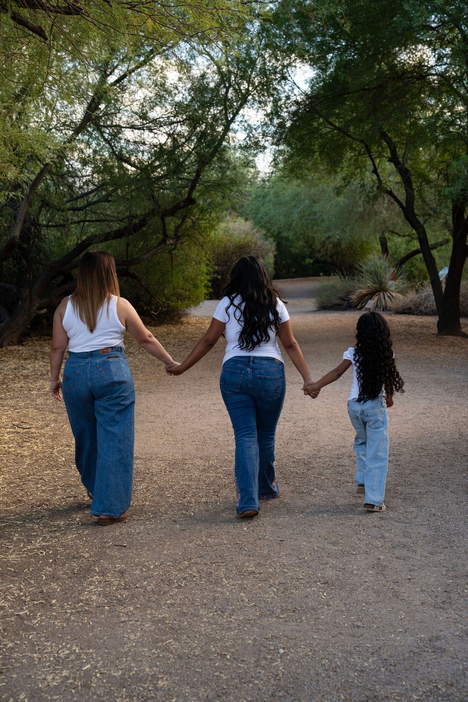 Two women and a young girl hold hands walking down a tree-lined path, all wearing white tops and jeans.